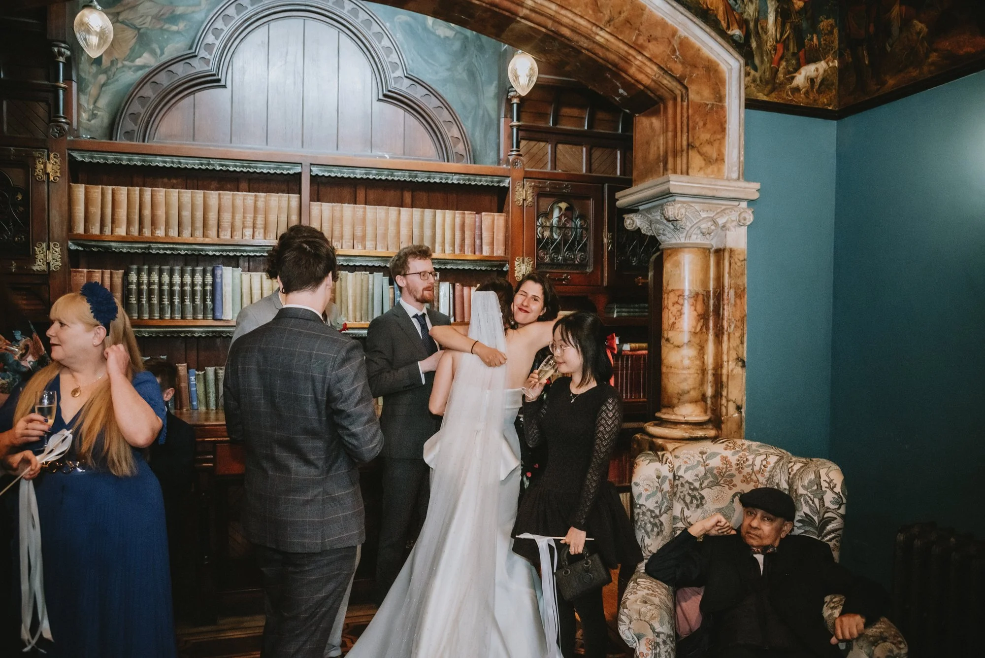 A wedding reception with a bride in a white gown holding a bouquet, surrounded by guests in formal attire, inside a room with a large bookshelf, ornate fireplace, and a teal wall.