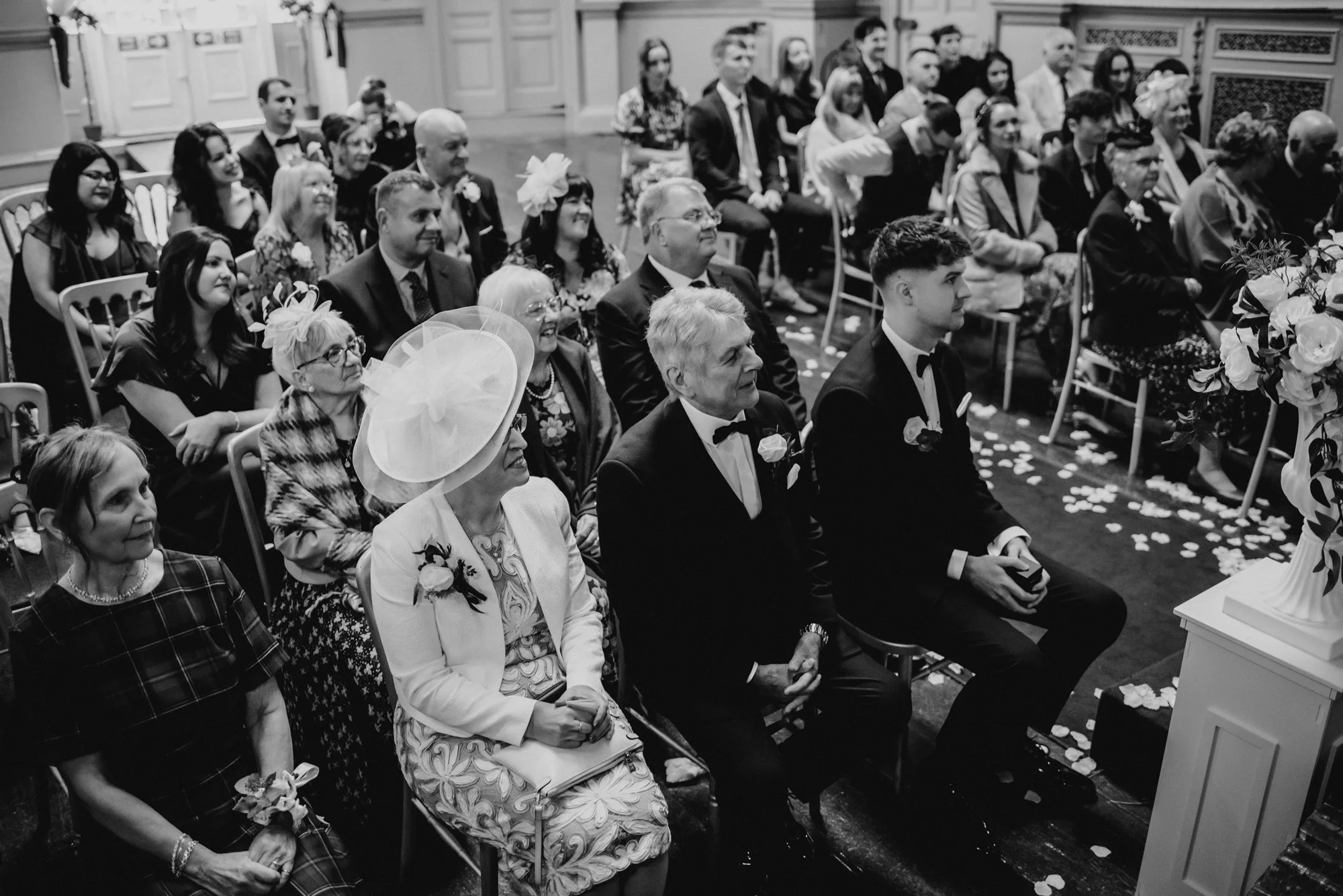 Guests seated in a wedding ceremony, including an elderly woman with a large hat, a man and a young man in tuxedos, and elegantly dressed women, with flower petals on the floor.