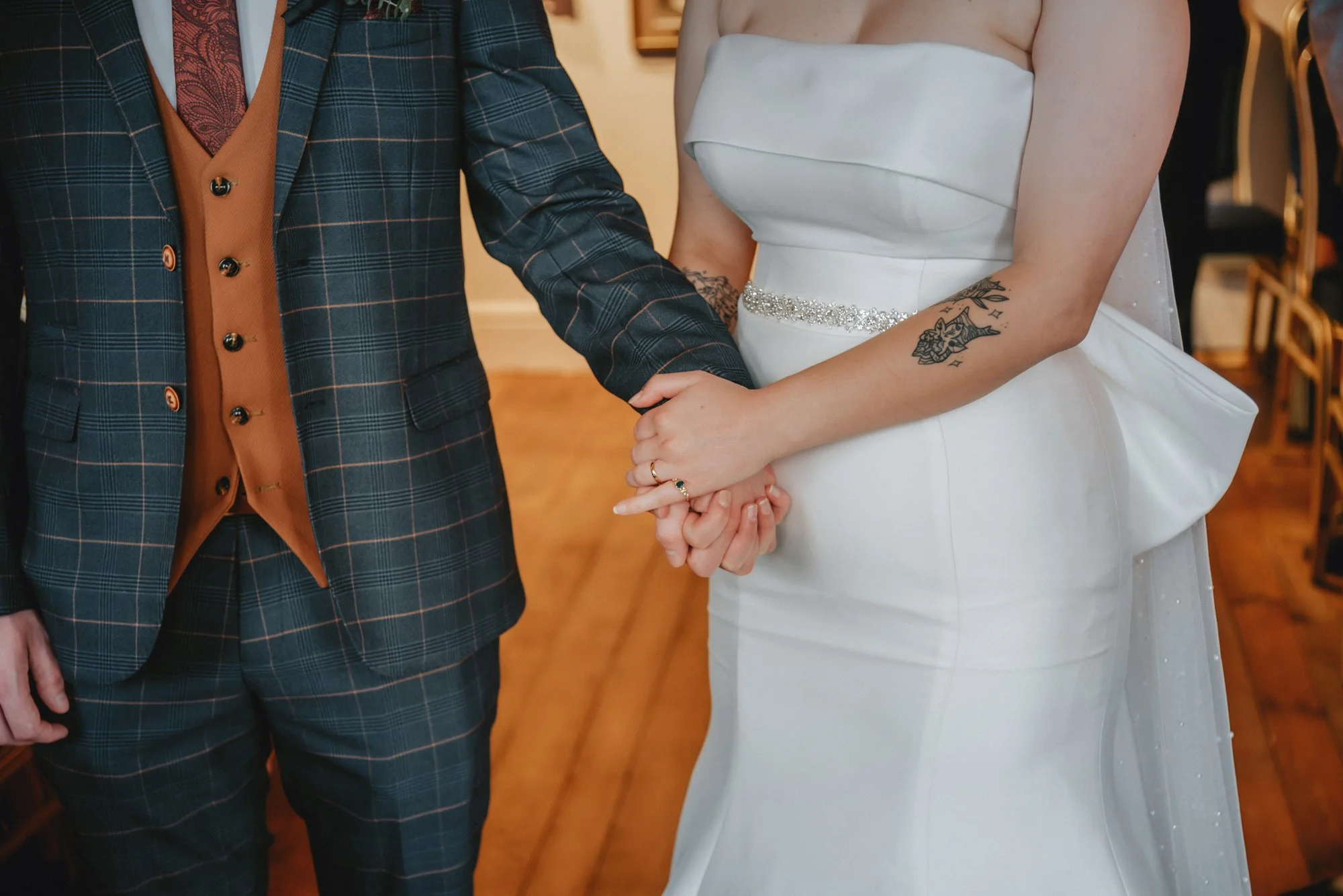 A bride and groom holding hands during their wedding ceremony, with the bride wearing a white strapless gown and the groom in a plaid suit with a brown vest.