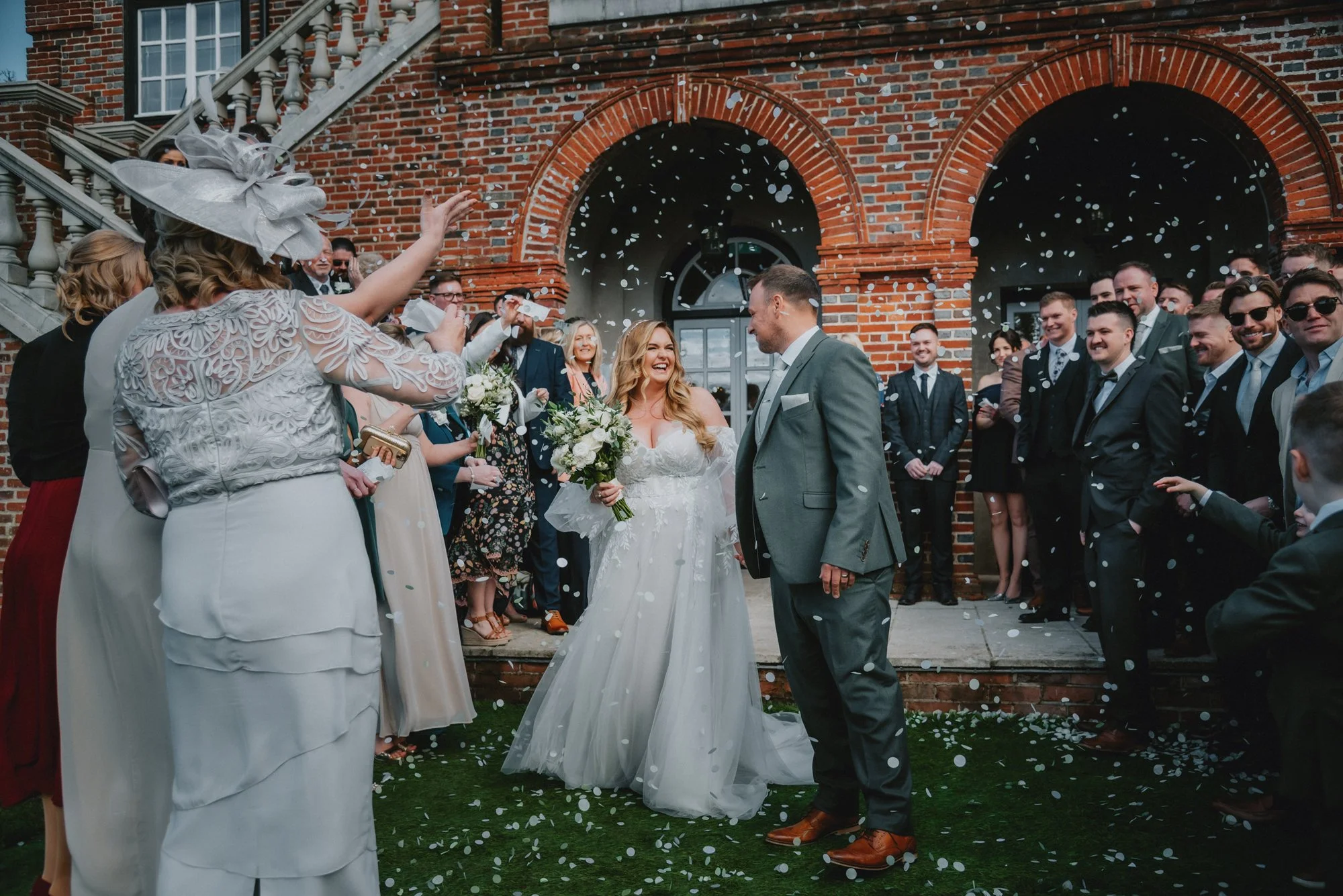 Bride and groom smiling at each other during wedding celebration outside a brick building, surrounded by guests throwing confetti.