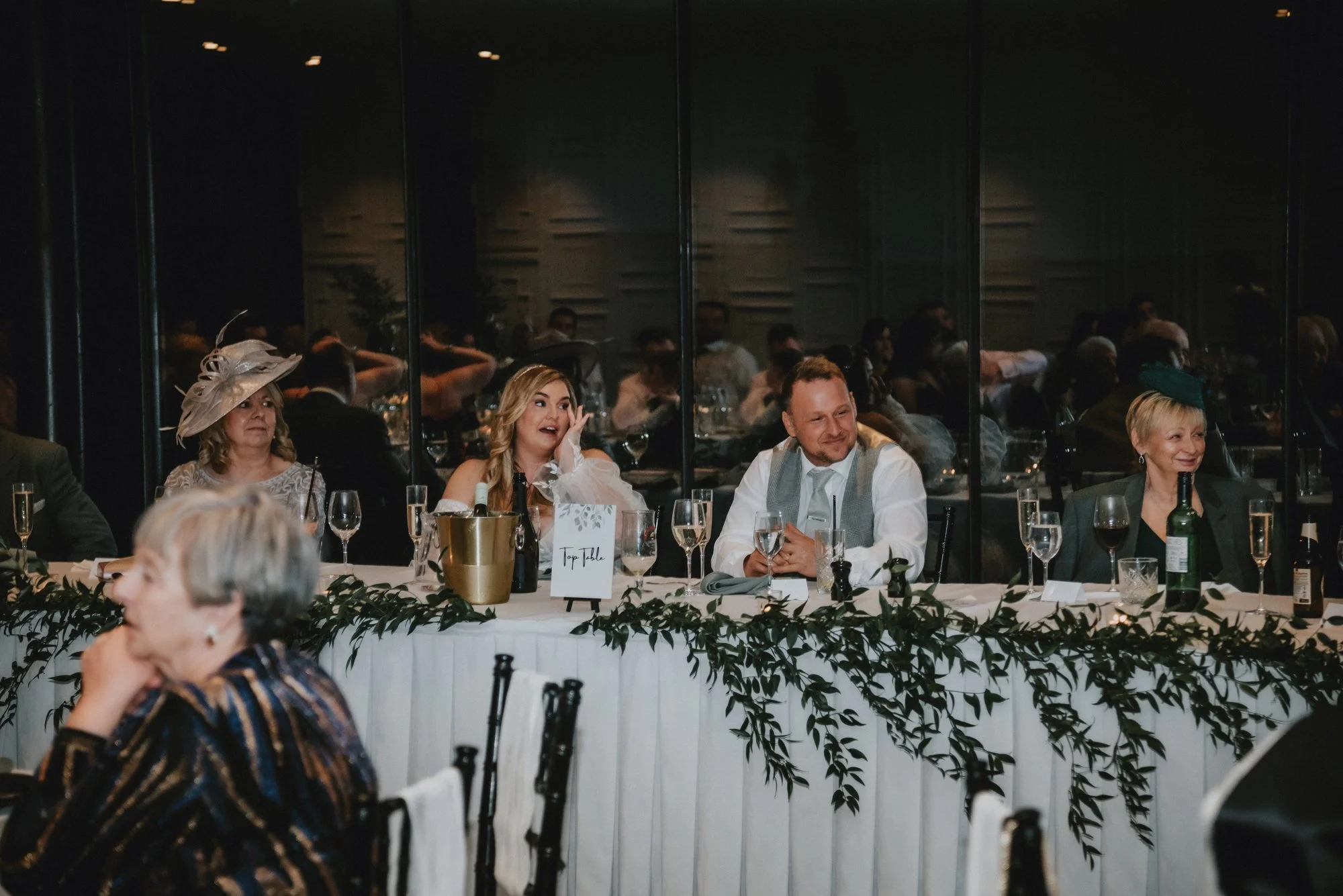 People sitting at a long decorated table during a formal event or wedding reception, with glasses and bottles, and a mirror reflecting the scene behind them.