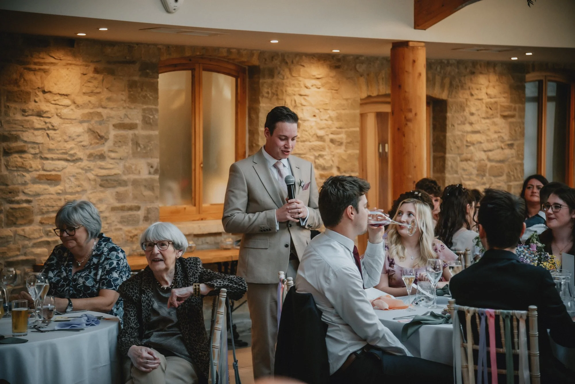 A man giving a toast at a wedding reception while another man drinks from his glass. The event is indoors with a rustic brick wall background and several guests seated at round tables.