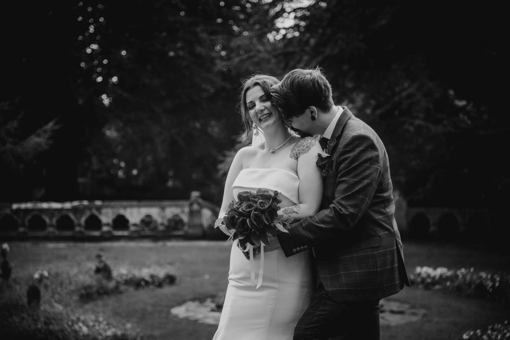 A black and white photo of a bride holding a bouquet of roses and a groom in a suit, embraced and smiling outdoors.