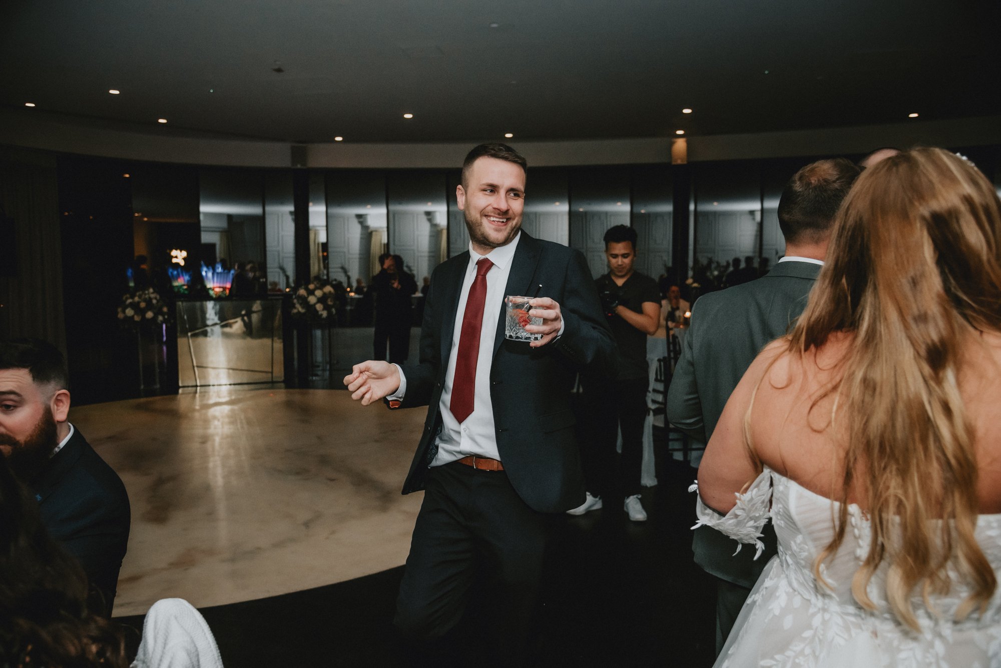 A man in a black suit, white shirt, and red tie smiling and holding a drink at a formal event or wedding reception, surrounded by other guests in formal attire.
