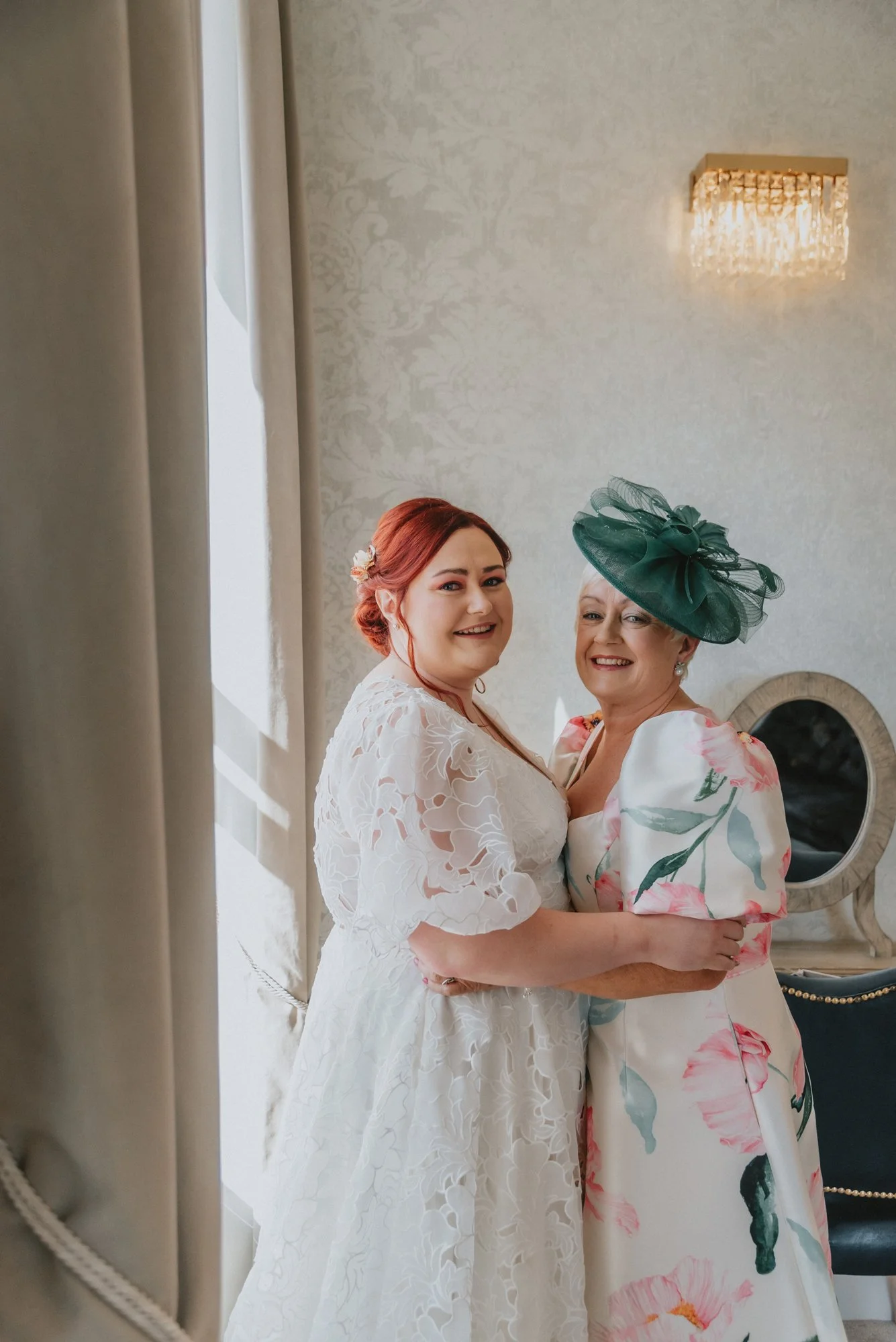 A bride and an older woman, possibly her mother, smiling and embracing each other indoors. The bride is wearing a white lace wedding dress, and the older woman is dressed in a floral dress with a large green hat. The setting appears to be a decorated