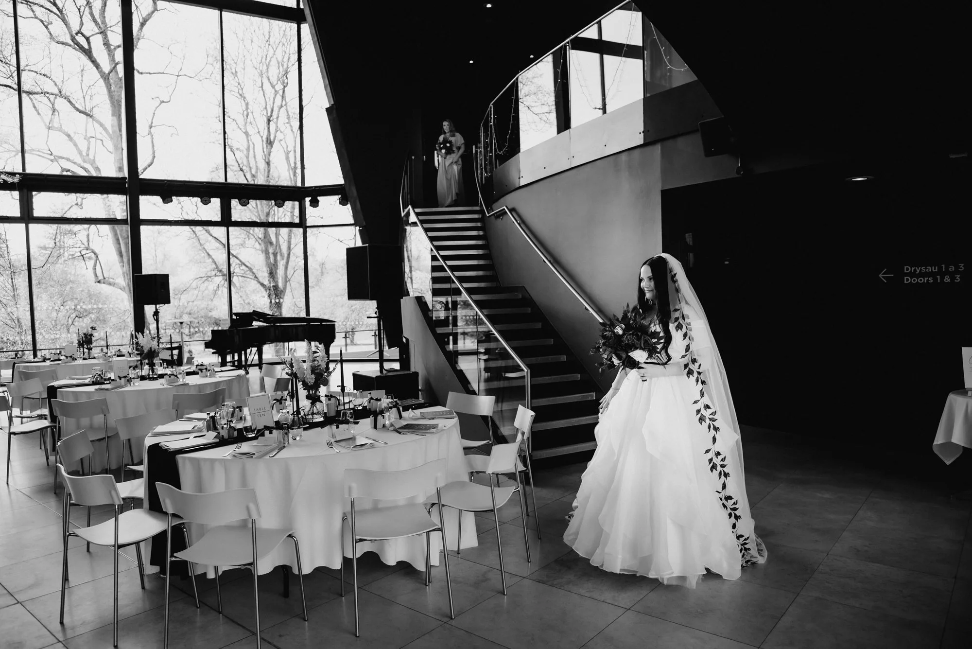 Black and white photo of a bride in a wedding dress holding a bouquet, walking near a staircase inside a modern venue with large windows showing trees outside, decorated round tables set for a wedding reception.