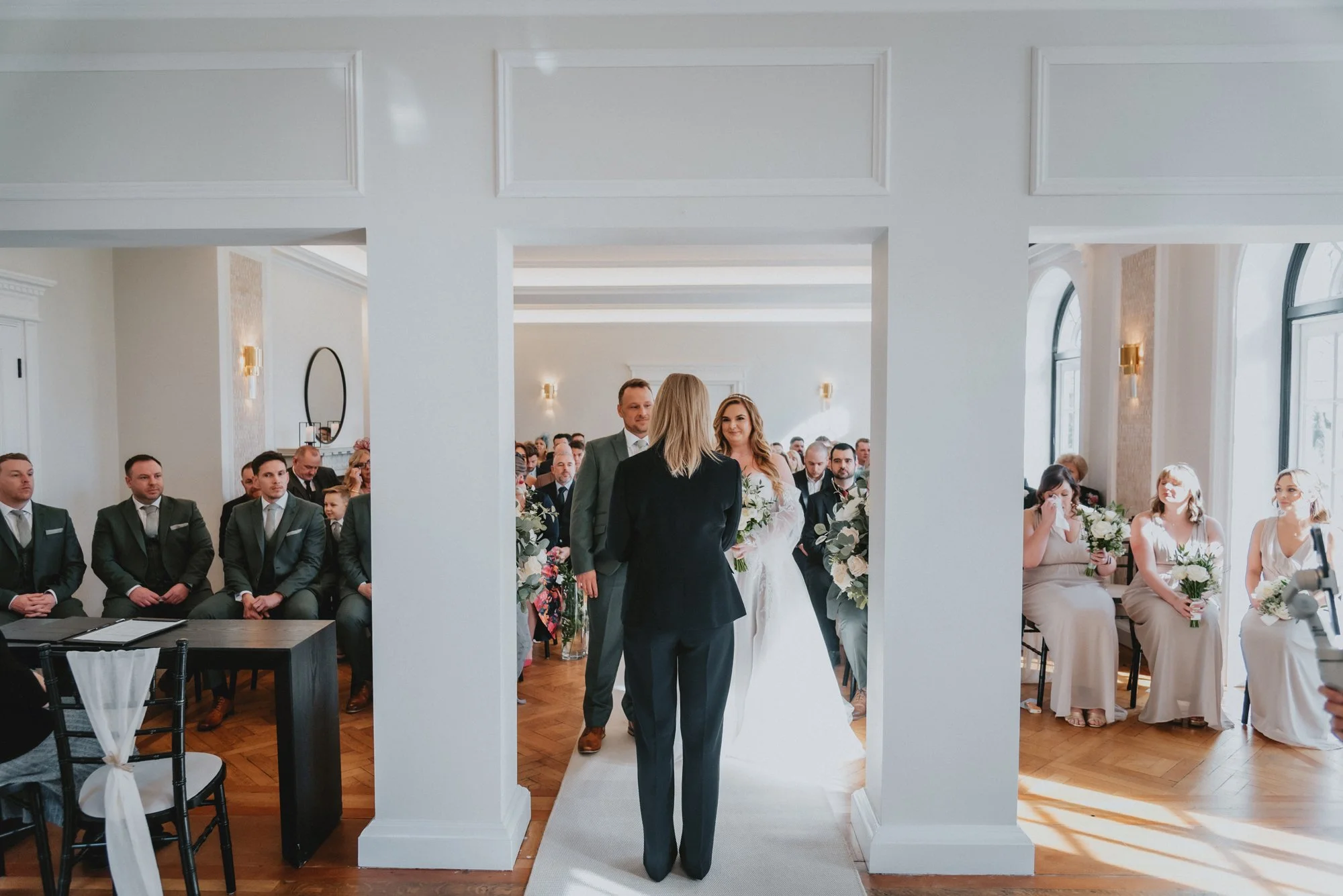A wedding ceremony with a bride and groom exchanging vows in front of seated guests in a bright, elegant indoor setting.
