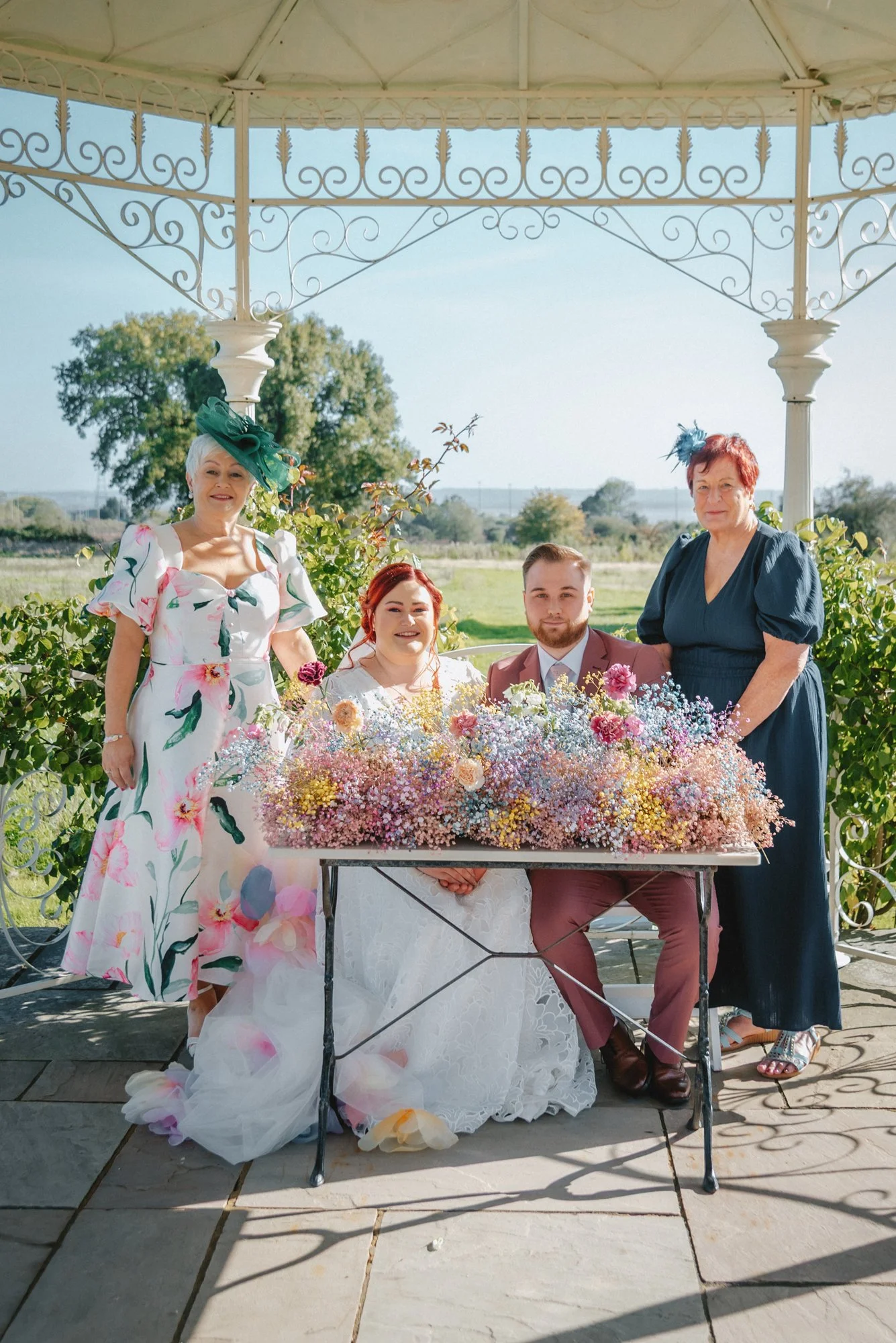 Group of four people celebrating a wedding outdoors with a scenic landscape in the background, featuring a bride in a white lace gown sitting at a table with colorful flowers, a groom in a pink suit sitting next to her, and two women standing on eith