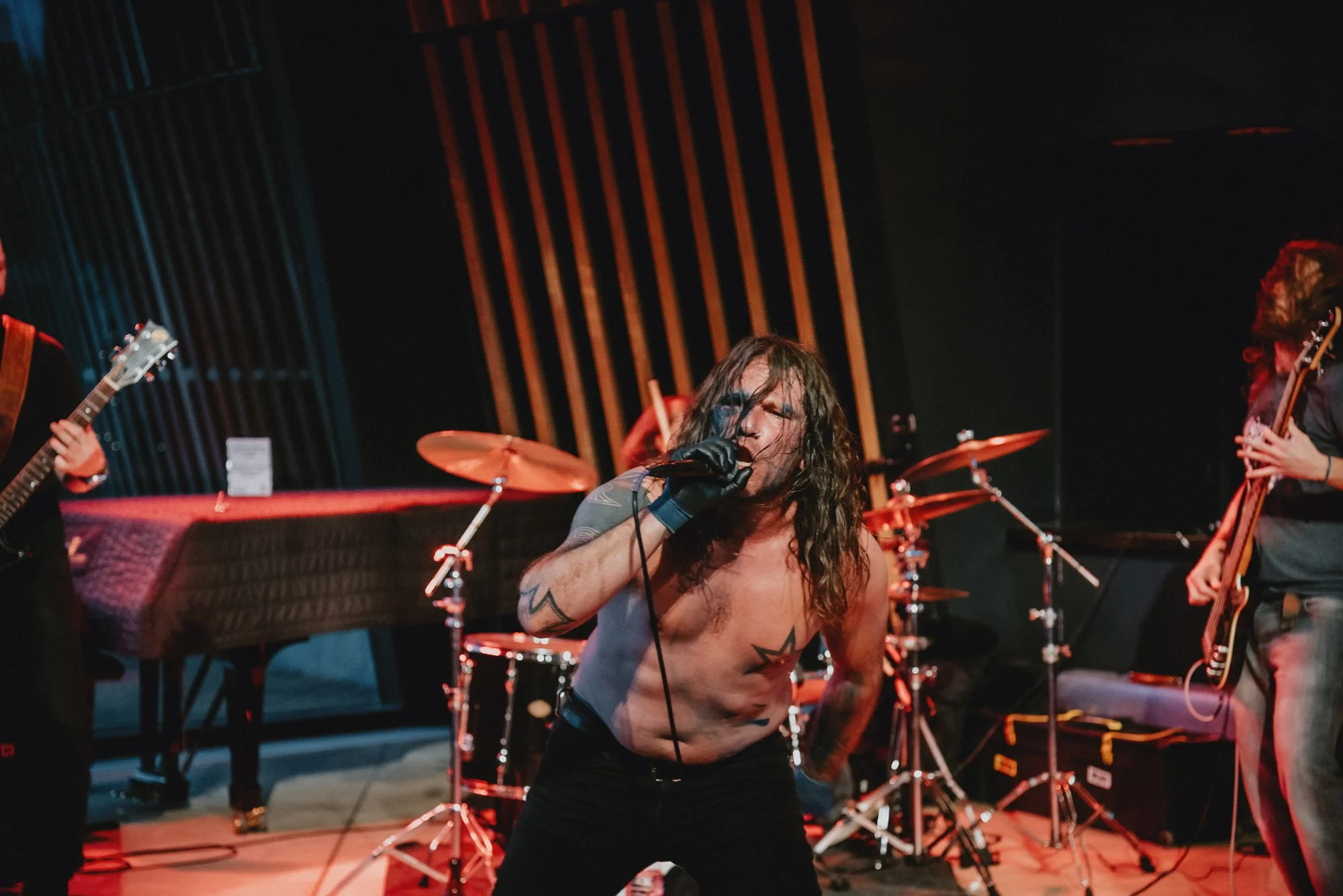A shirtless male lead singer with long hair and tattoos singing into a microphone on stage during a rock concert, with a band playing instruments behind him.