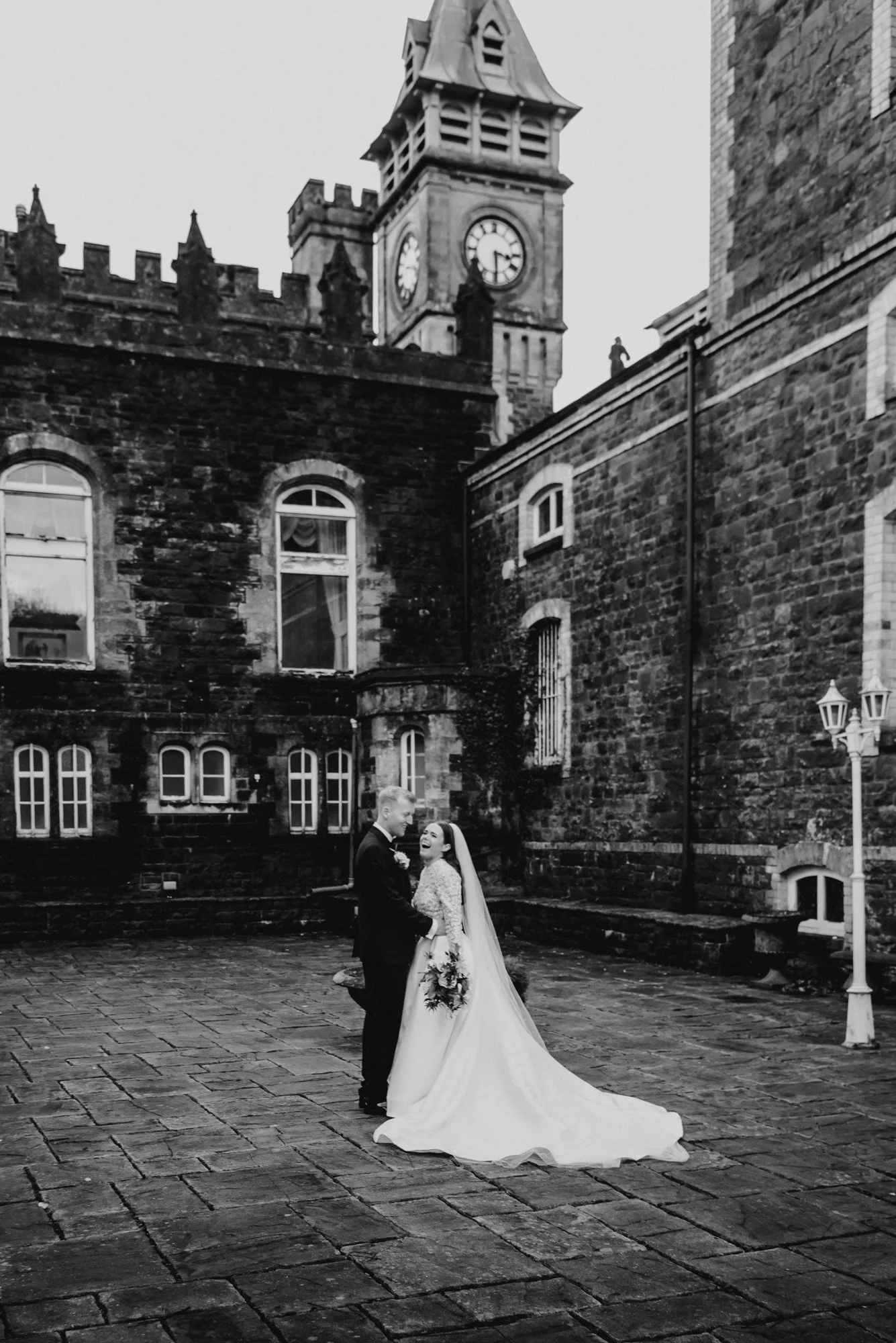 A black and white photo of a bride and groom in wedding attire standing on cobblestone ground in front of a historic brick building with a clock tower, smiling and holding hands.
