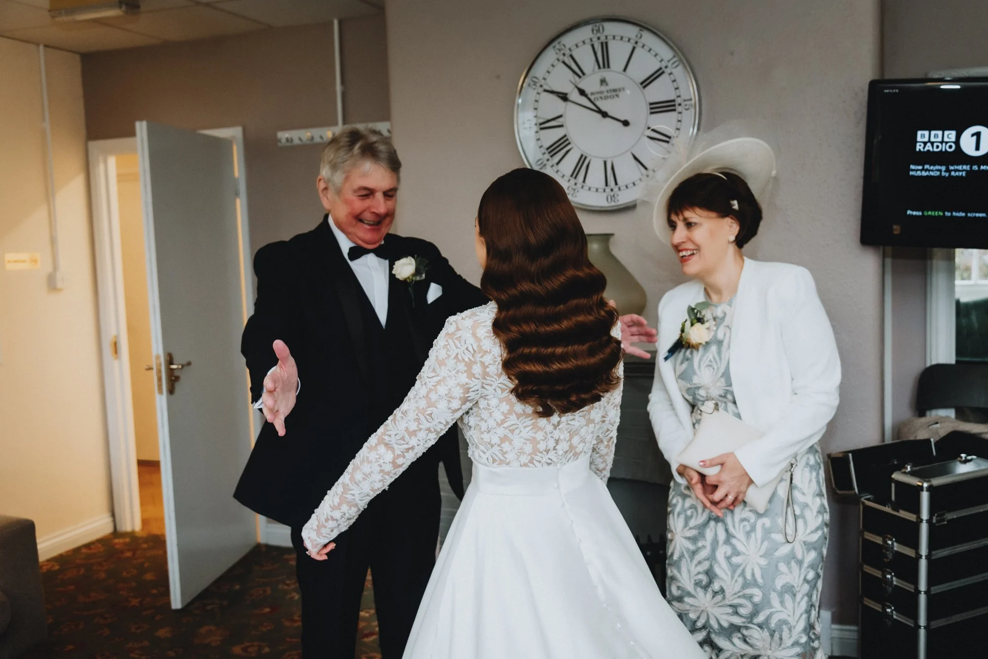 A bride in a white wedding gown greeting a man in a tuxedo, with a woman in a white dress and hat smiling nearby, inside a room with a large wall clock and TV.