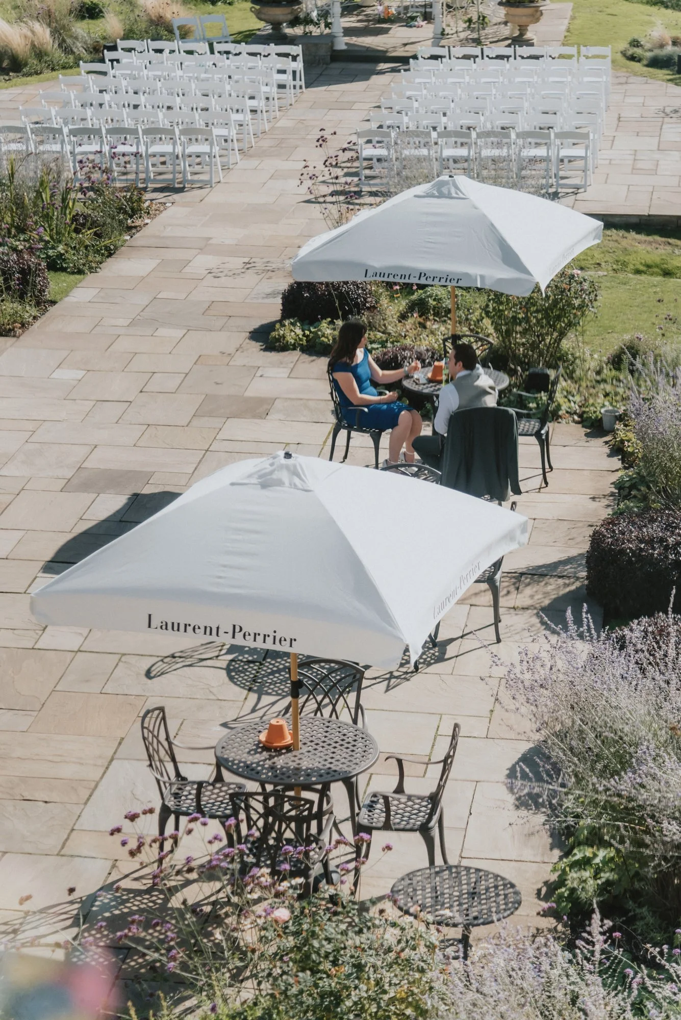 A sunny outdoor patio with white chairs set up for a wedding ceremony, two tables with chairs and large white Laurent-Perrier branded umbrellas, and a couple sitting at a small table under one umbrella, engaged in conversation.