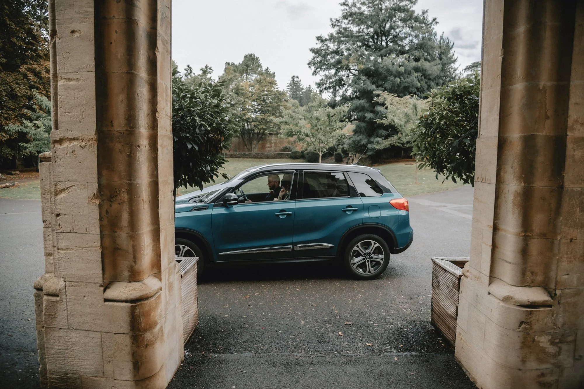 View of a blue SUV parked outside between stone columns with a man sitting inside, surrounded by a lush green landscape with trees and grass.