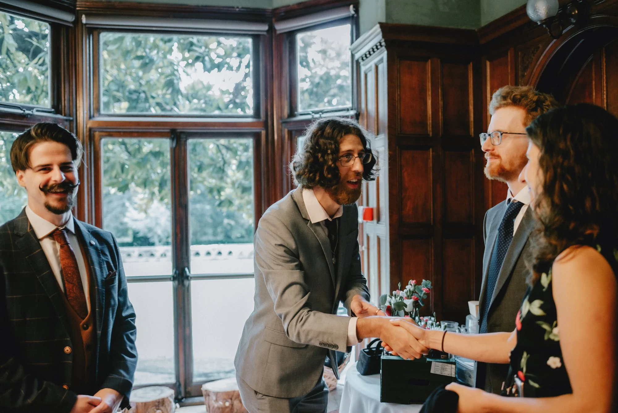 A man with curly hair and glasses shaking hands with a woman at an indoor event, with two men standing nearby, in front of large windows and wooden paneling.