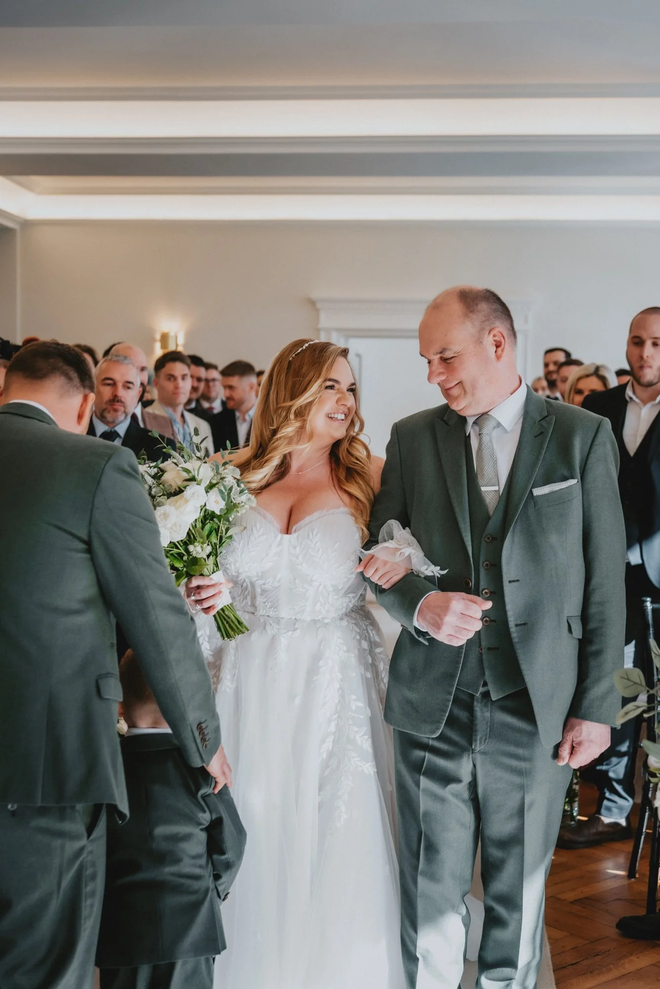 A bride in a white wedding dress smiling at her father during a wedding ceremony, holding a bouquet of white flowers. The groom is also visible, standing to the left.