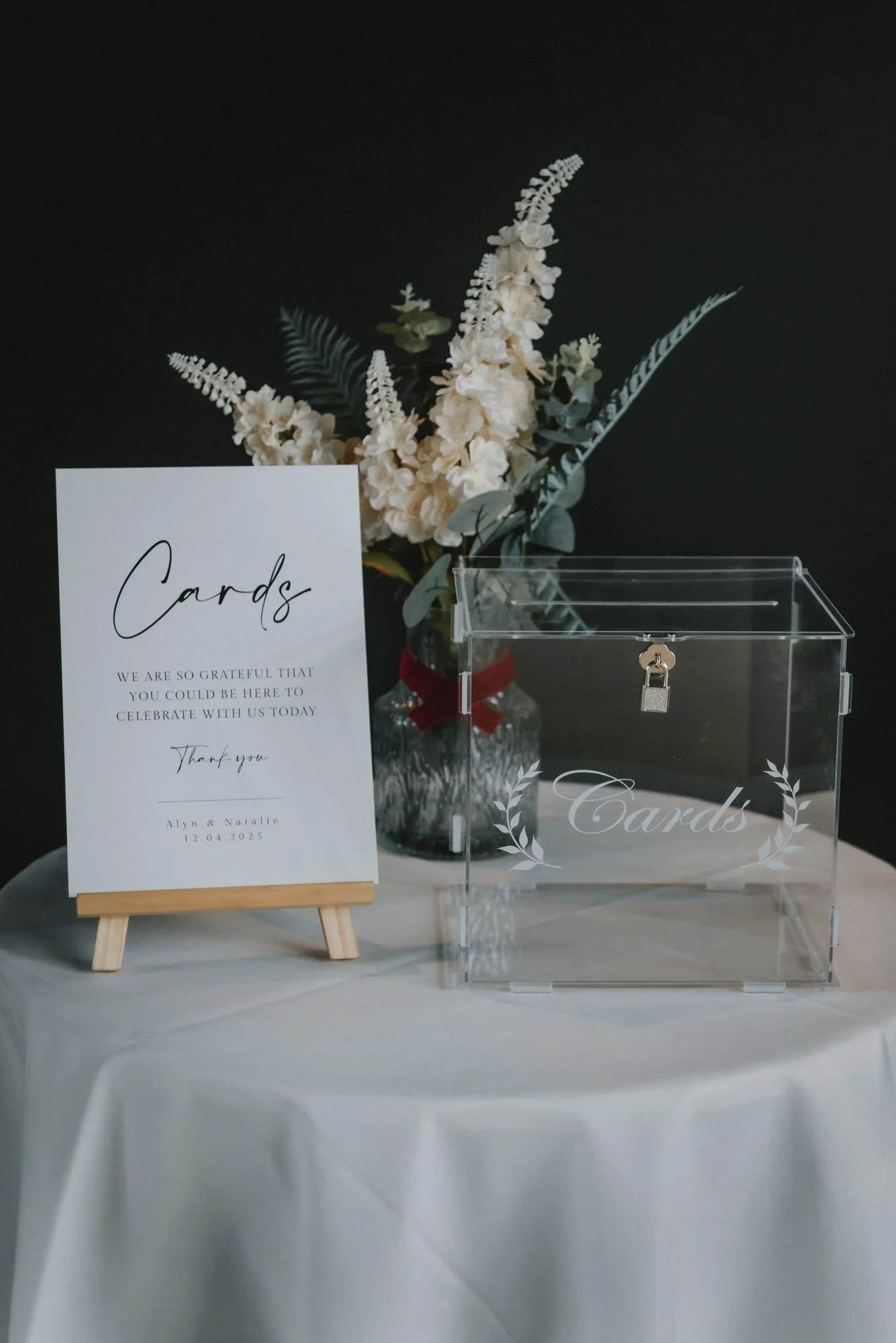 A clear acrylic card donation box with the word 'Cards' and a decorative laurel design written on it, placed on a white table. Next to it, a white sign on a small easel with the word 'Cards' in cursive, a message of gratitude, and the date 12.04.2025