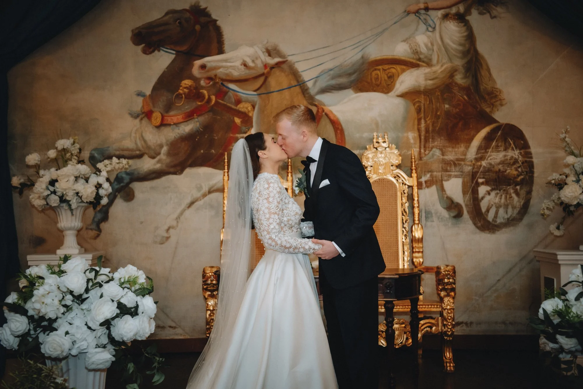 A bride and groom kiss during their wedding ceremony, standing in front of a large mural of a chariot pulled by horses.