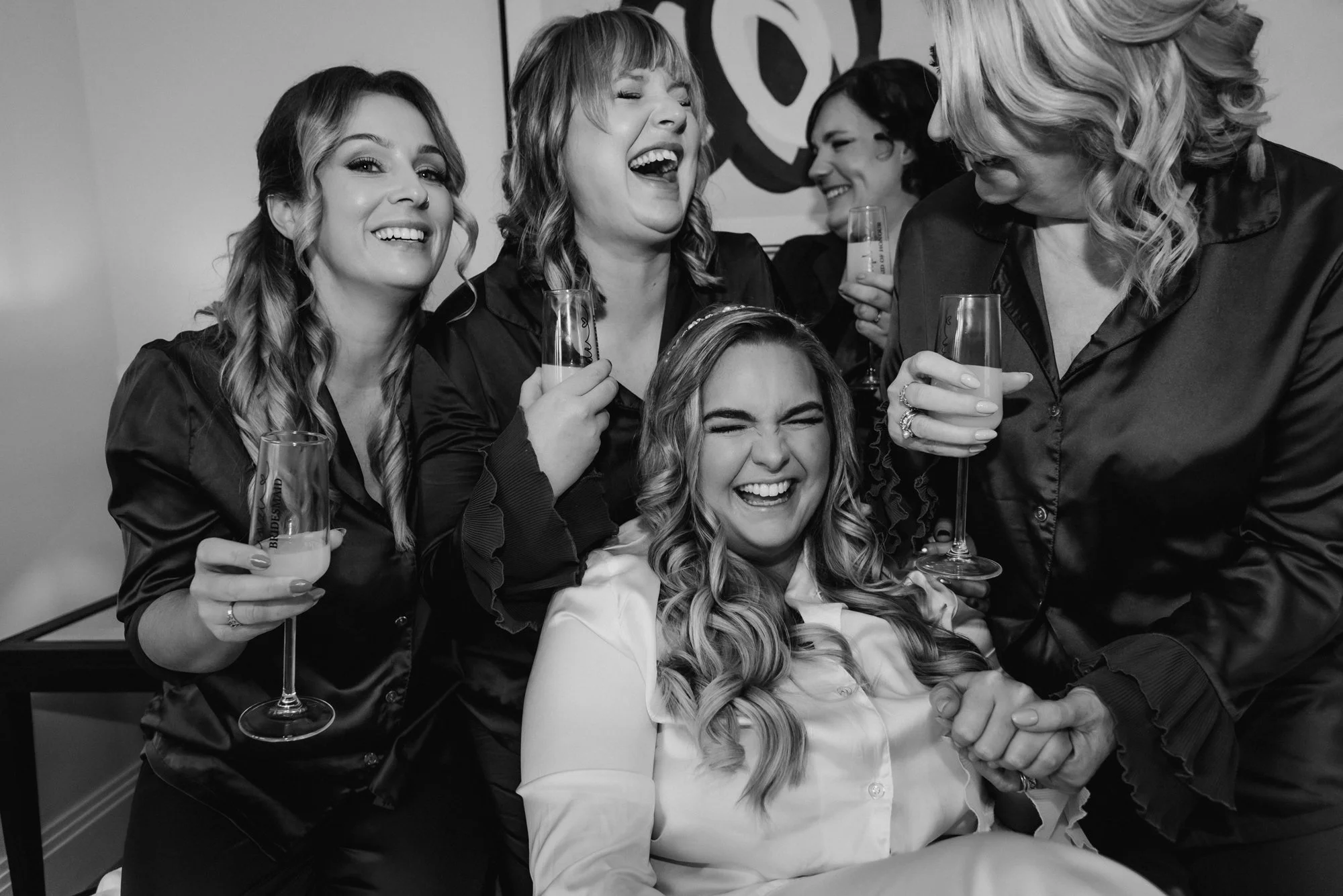 Group of five women celebrating with drinks, laughing, and smiling together in a cozy indoor setting.