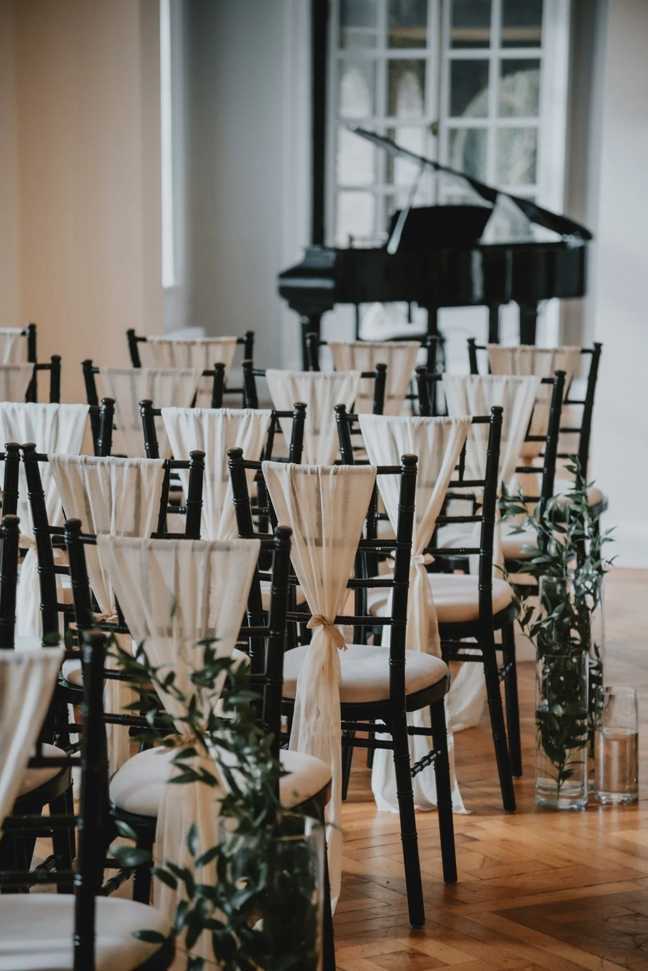Rows of black chairs with cream-colored fabric drapes tied on the back, arranged in an indoor event space with a piano and large windows in the background.