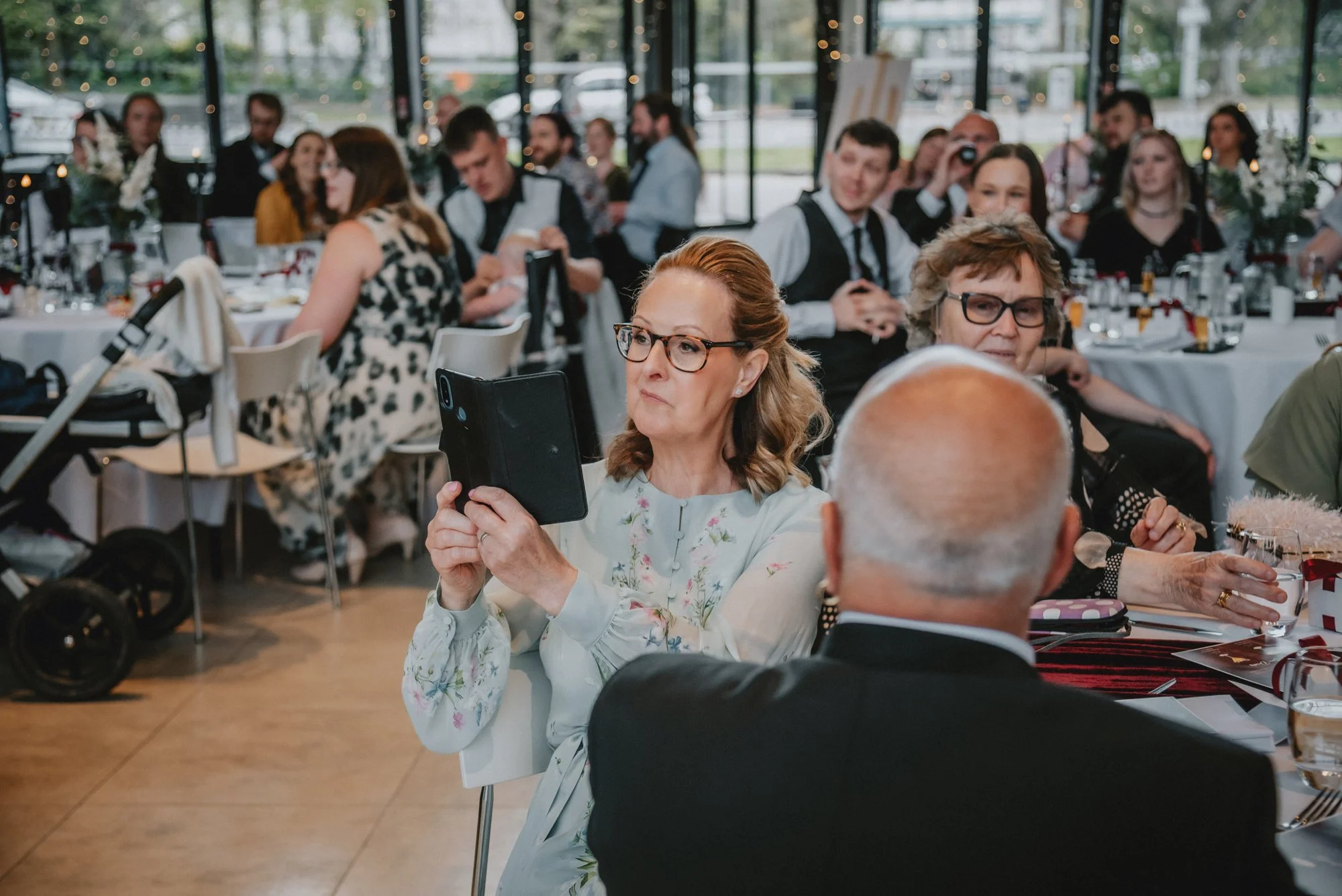A woman with glasses and floral blouse sitting at a table, holding a tablet with both hands, at a formal event or wedding reception. Other guests are sitting at round tables in the background, some taking photos or looking around.