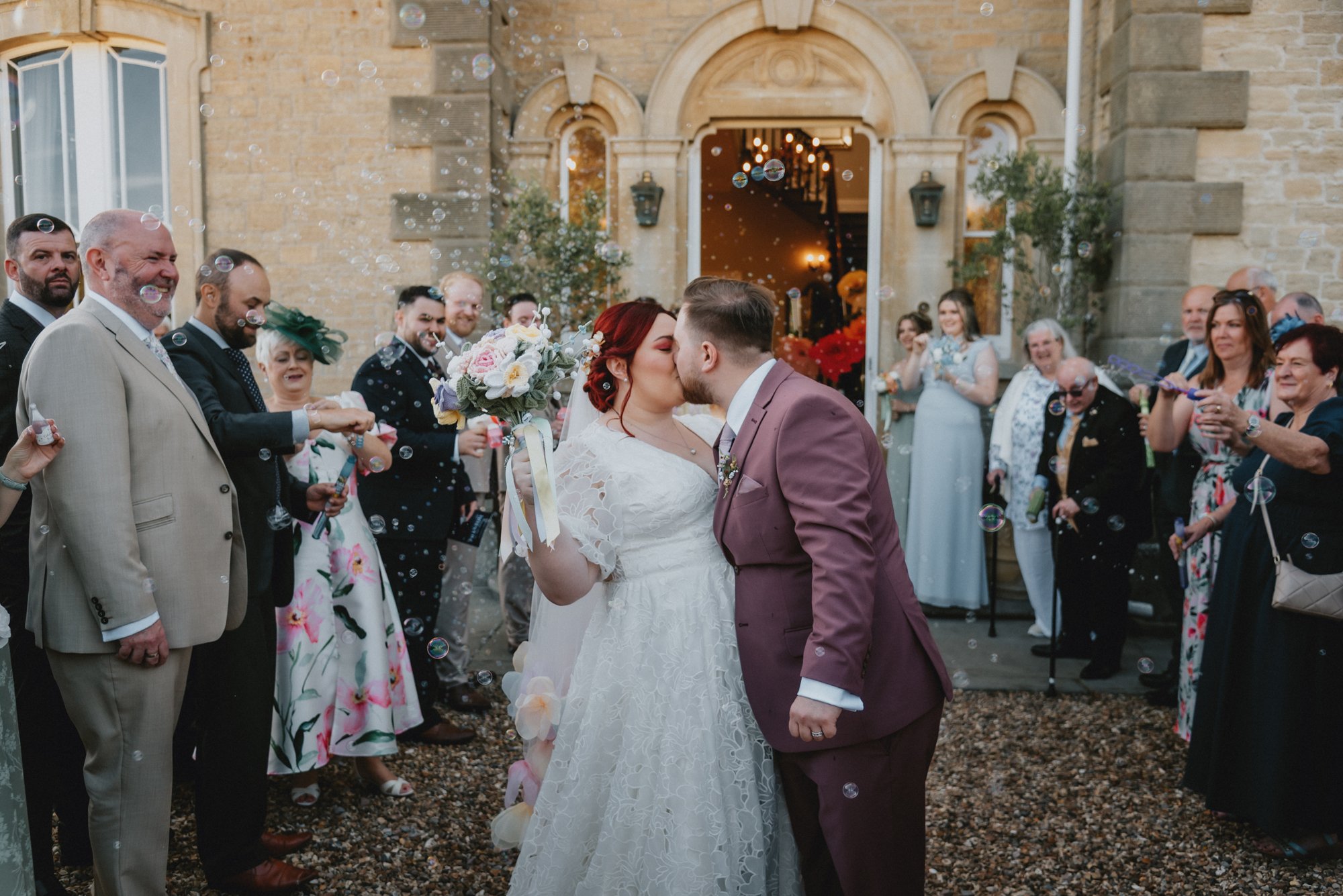 Bride and groom kiss outside a wedding venue surrounded by guests celebrating with bubbles.
