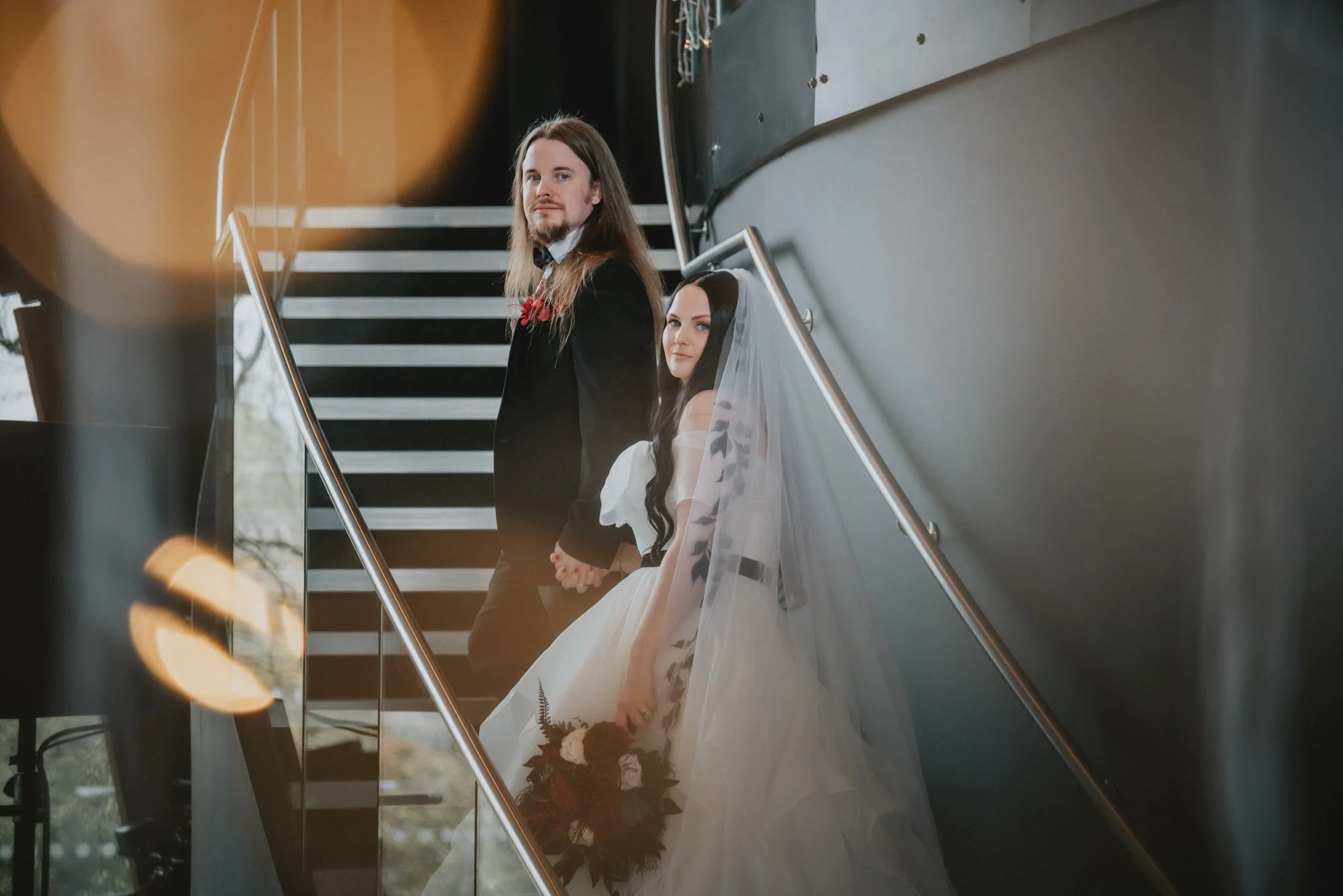 A bride and groom standing on a staircase, holding hands, dressed in wedding attire, with the bride holding a bouquet, in an indoor setting.