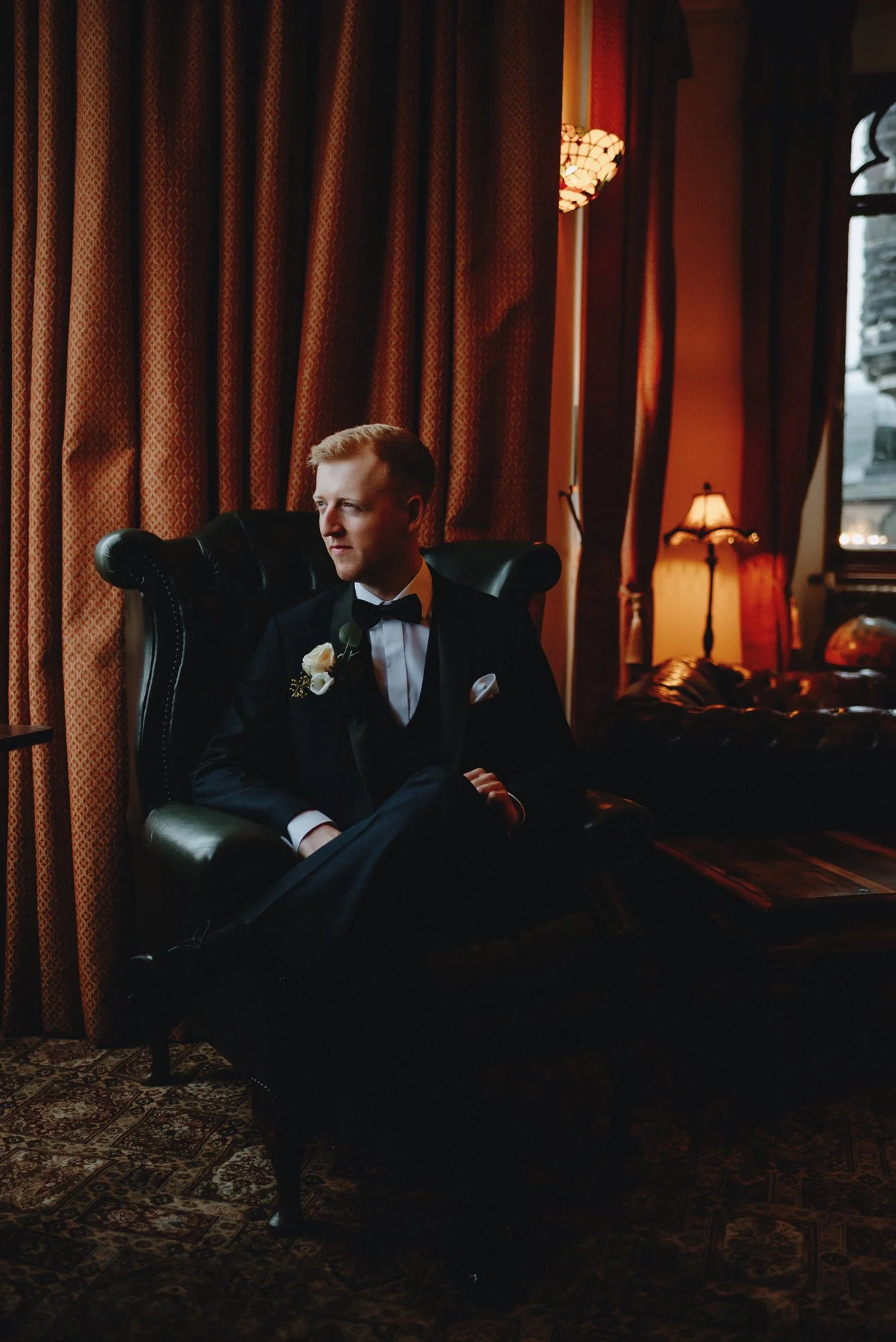 A man in a tuxedo sitting on a vintage black leather armchair in a dimly lit room with ornate curtains and warm lighting.