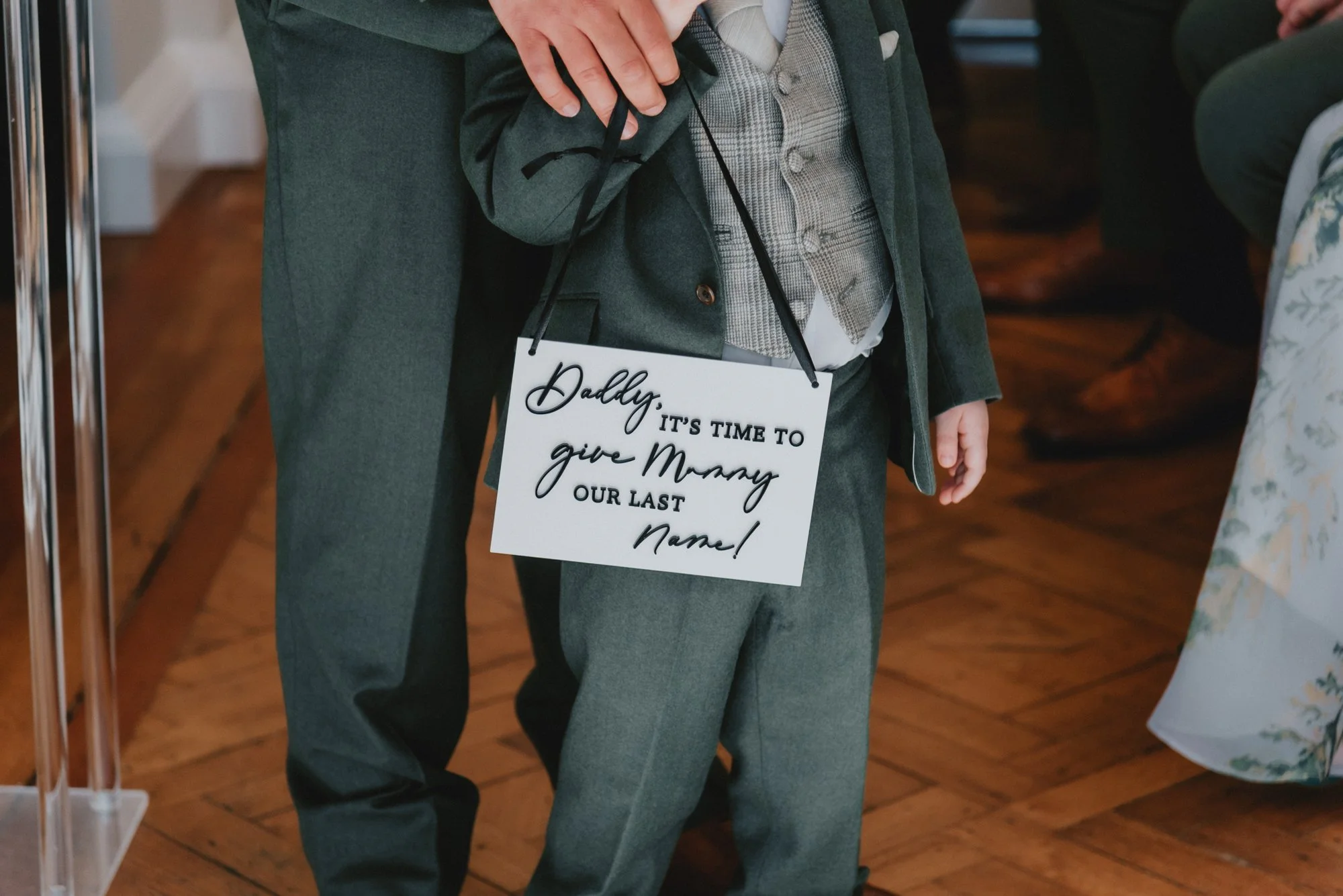 A young boy dressed in formal clothing wearing a sign around his neck that says: "Daddy, It's time to give Memy our last name!"