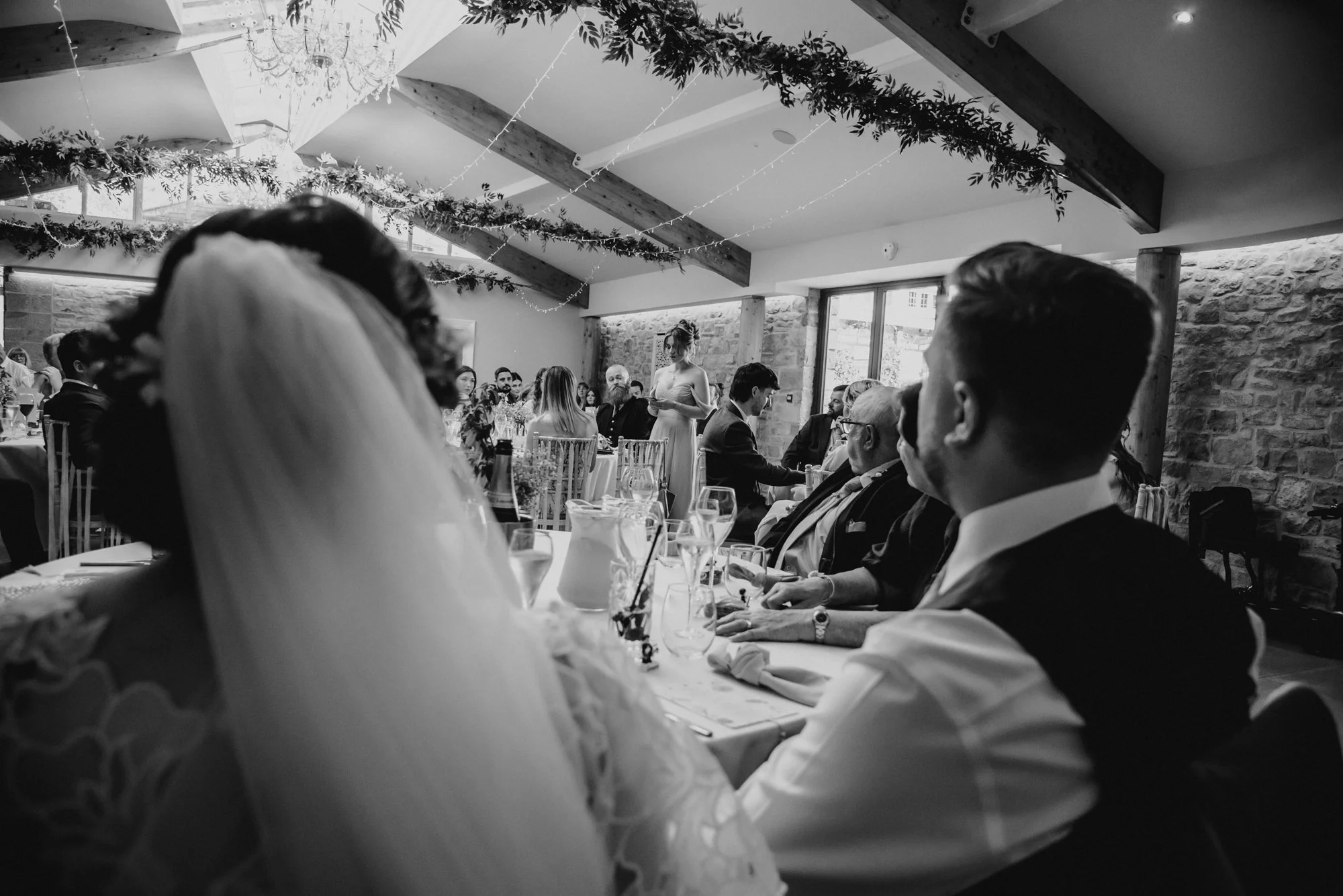 Black and white photo of a wedding reception, showing a bride with a veil and wedding dress in the foreground, seated at a long table with guests, with a woman in a formal dress standing and giving a speech in the background in a rustic venue with wo