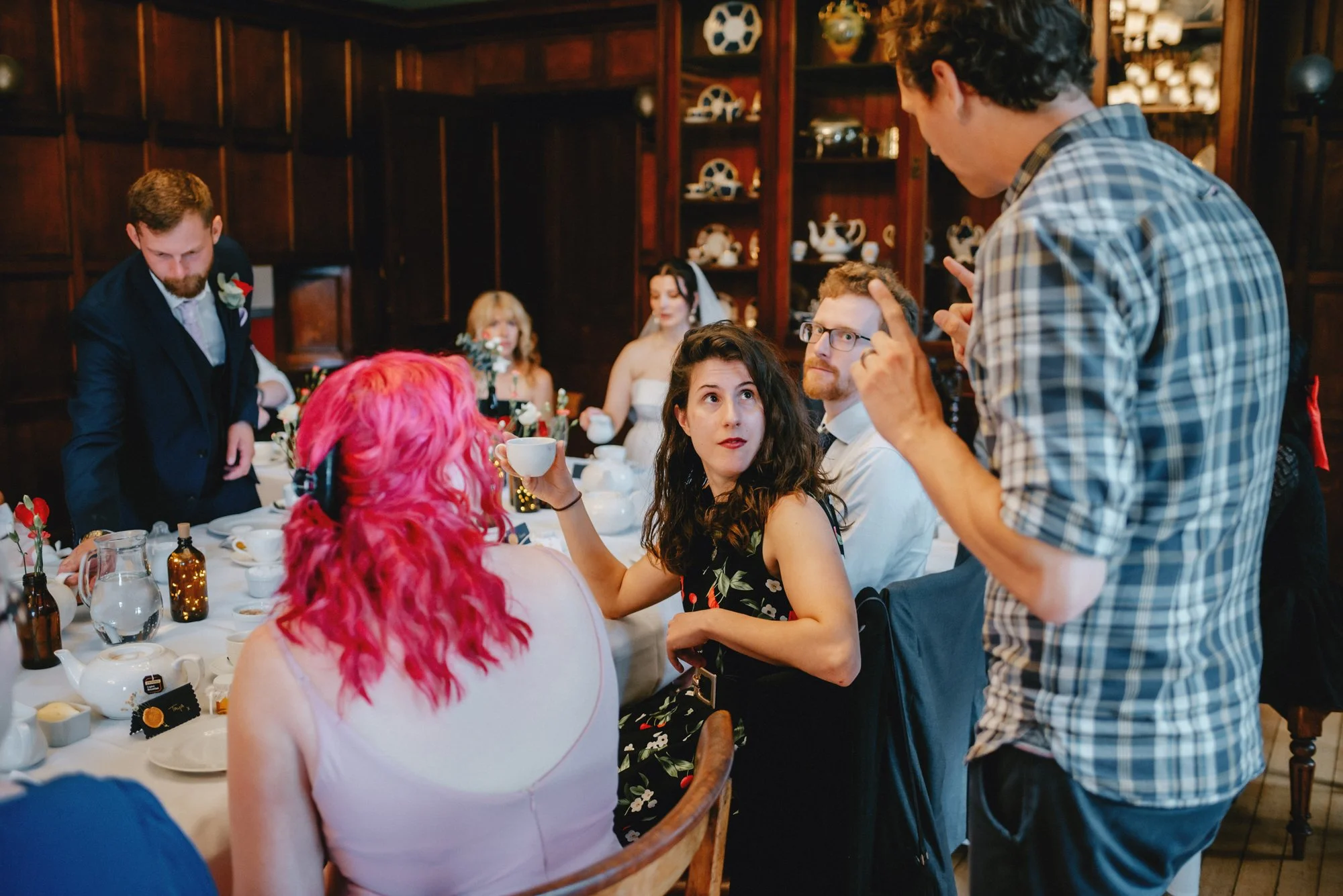 People seated at a dining table engaged in conversation during a gathering or celebration in a wood-paneled room with shelves of teapots and decorative items.