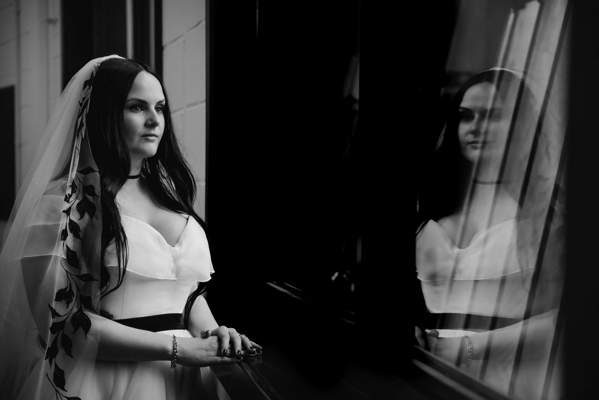 Black and white photo of a woman in a wedding dress and veil, standing beside a window, with her reflection visible on the glass.