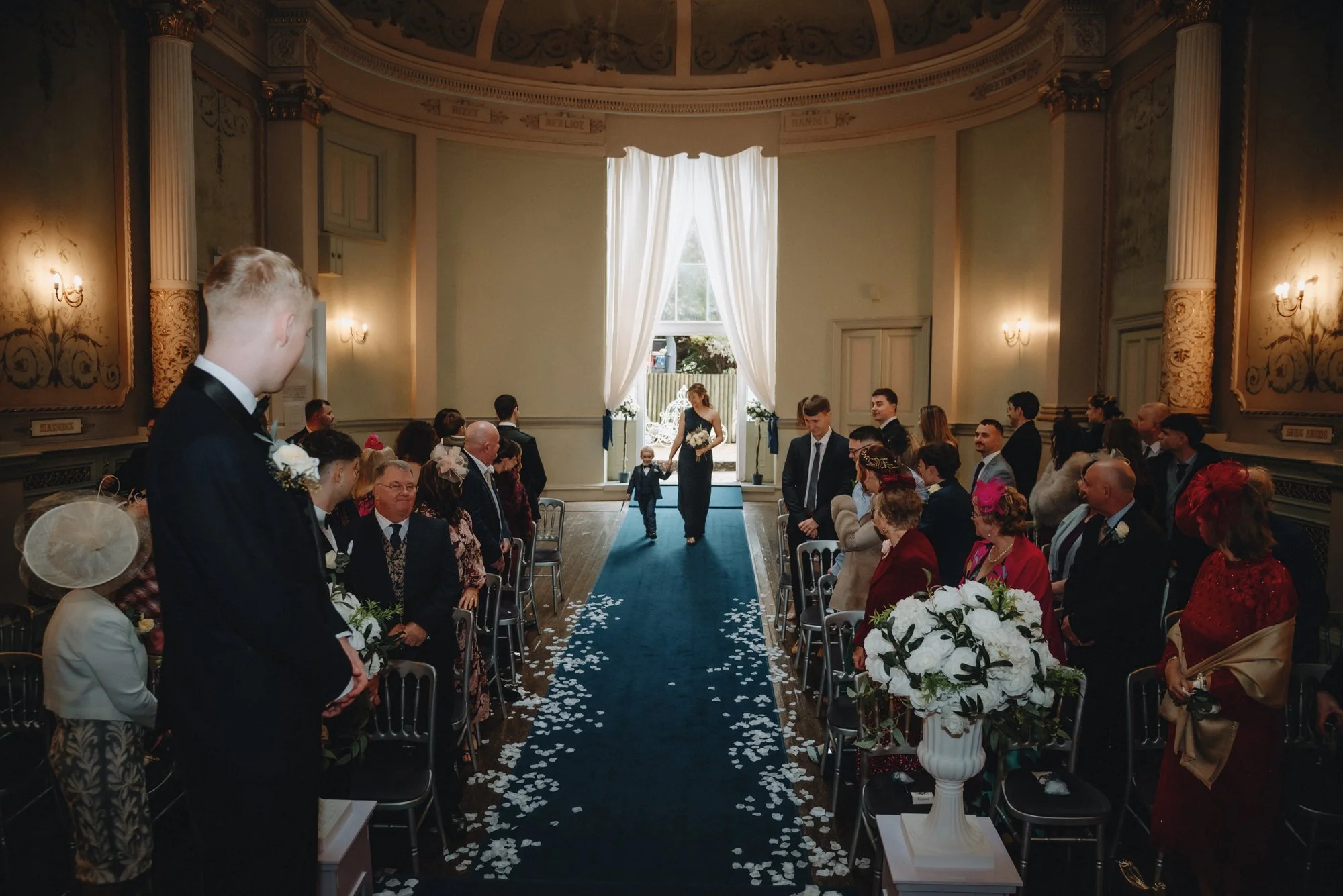 A wedding ceremony taking place in a historic church with high ceilings, ornate wall decorations, and large windows with white curtains. The bride, dressed in black, walks down the aisle with a child, while guests are seated on either side, some stan