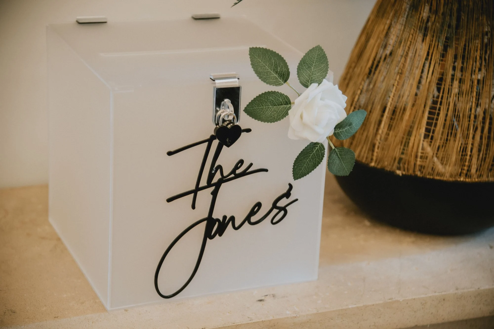 Decorative box with a white rose, green leaves, and a black script sign that says "The Jones", placed on a beige surface with a woven lamp in the background.