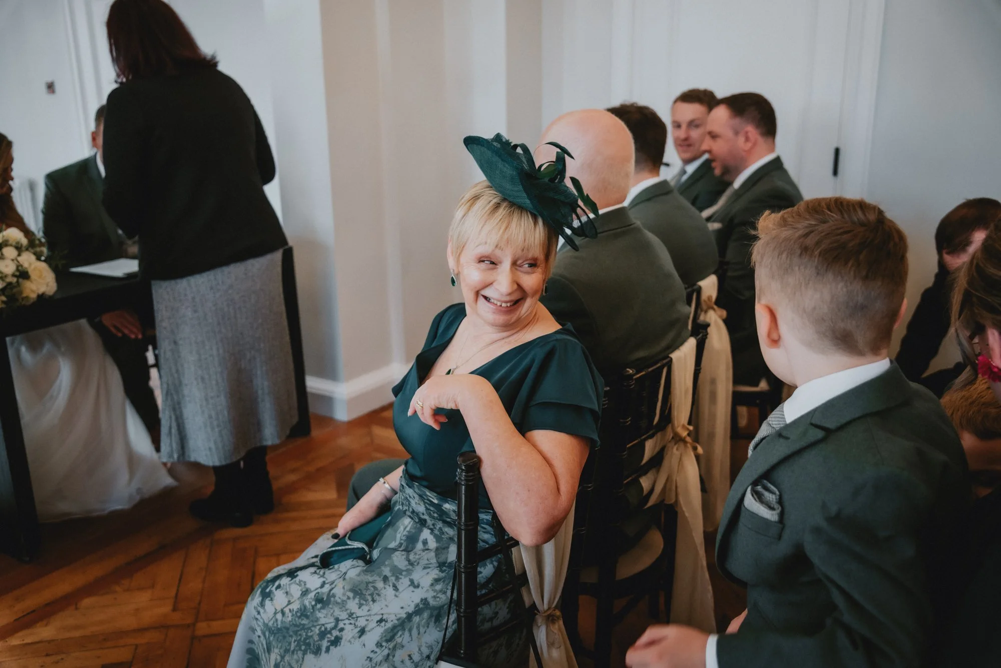 An elderly woman in a teal dress with a matching hat, sitting on a chair, smiling and looking over her shoulder at a young boy in a suit at a formal event or wedding.