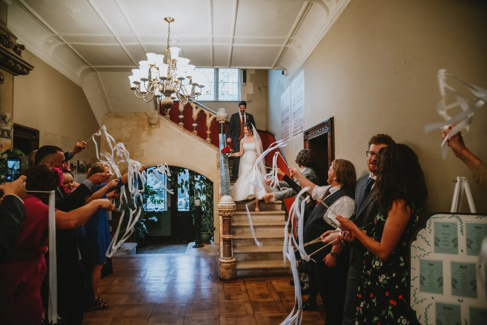 Bride and groom descending a staircase after a wedding ceremony, surrounded by guests throwing ribbons. The room features a chandelier, staircase with red carpet, and vintage decor.