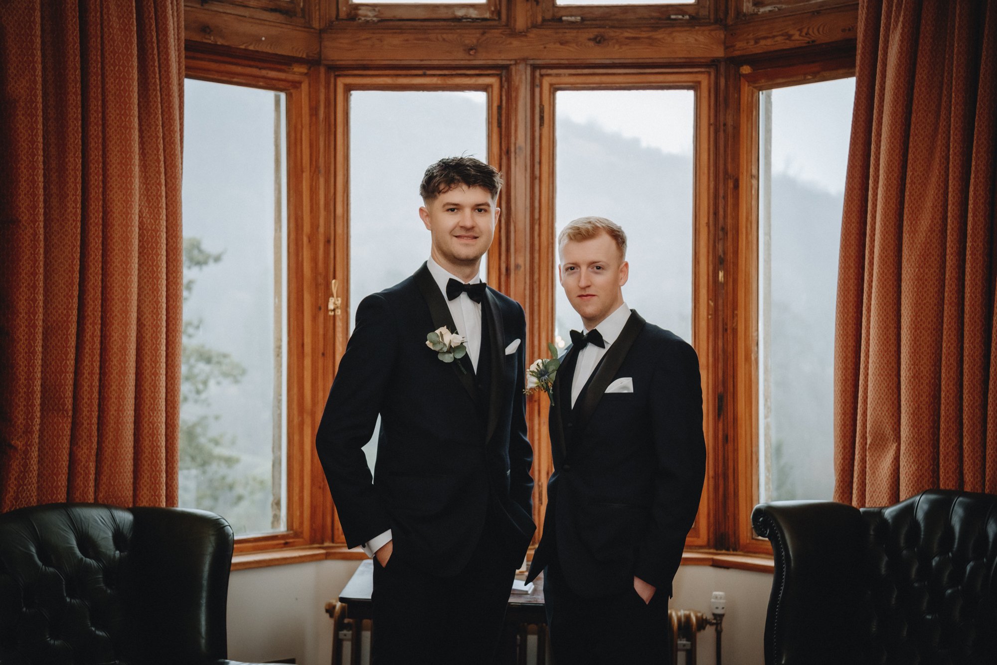 Two men in tuxedos with bow ties and boutonnières standing in front of large wooden windows, smiling at the camera in a room with black leather chairs and curtains.