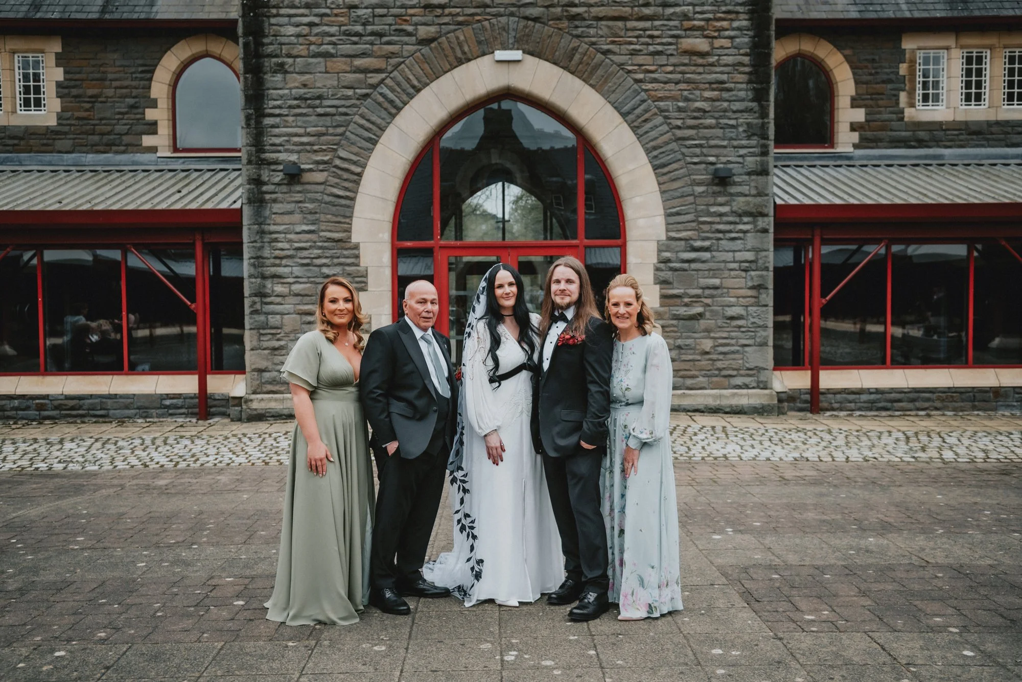 A group of five people gathered outside a stone building at a wedding, including a bride and groom in the center, flanked by two women and a man.