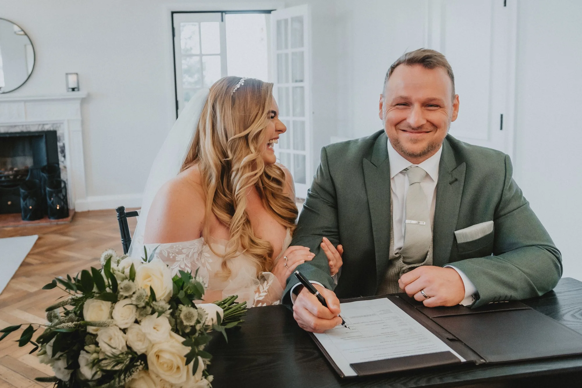 A bride and groom signing a marriage license at a wedding ceremony, with the bride smiling and looking at the groom, who is smiling and holding a pen over the document on a desk.