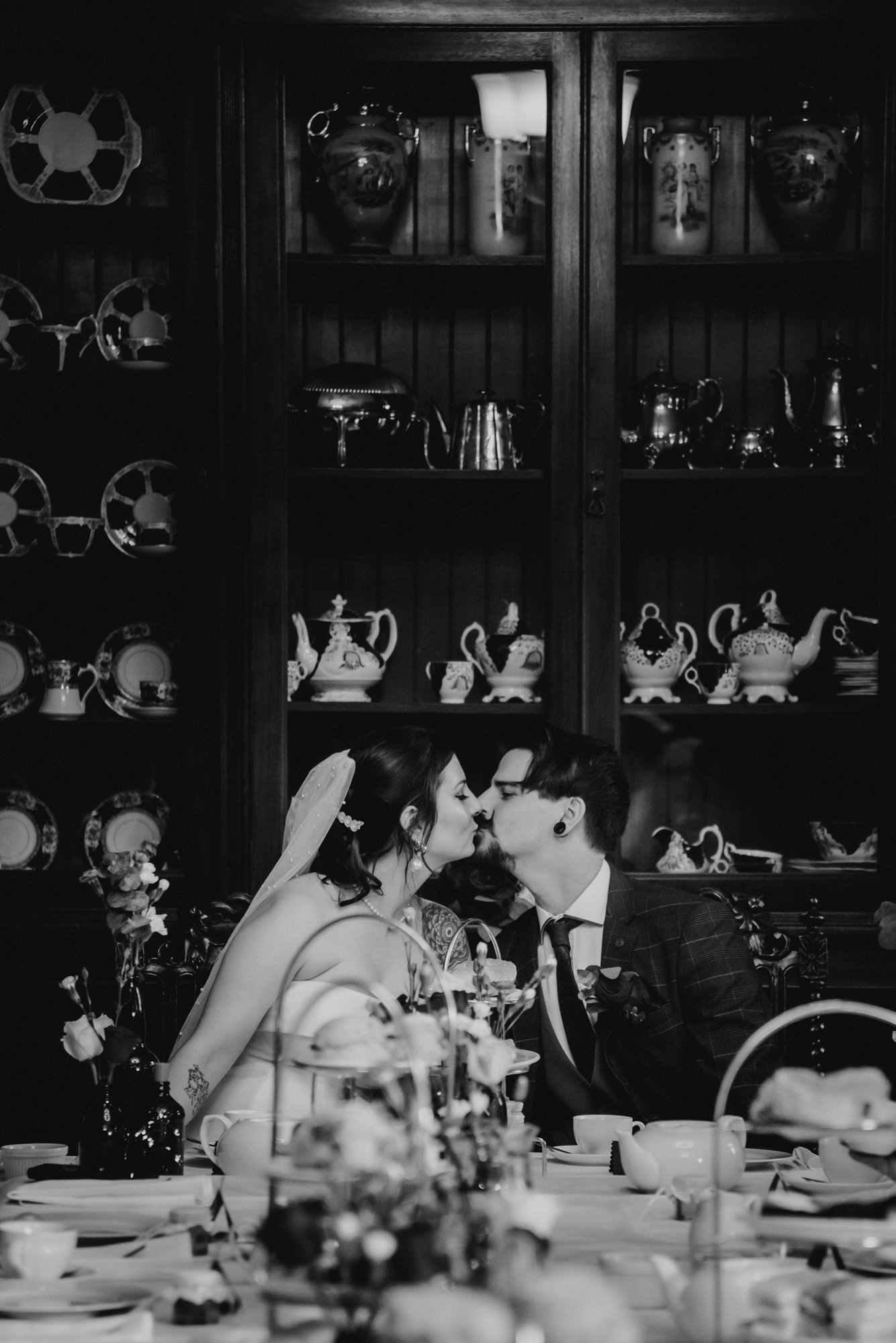 A black and white photograph of a bride and groom sharing a kiss at their wedding reception, sitting at a table with floral centerpieces, in front of a wooden cabinet filled with china and decorative teapots.