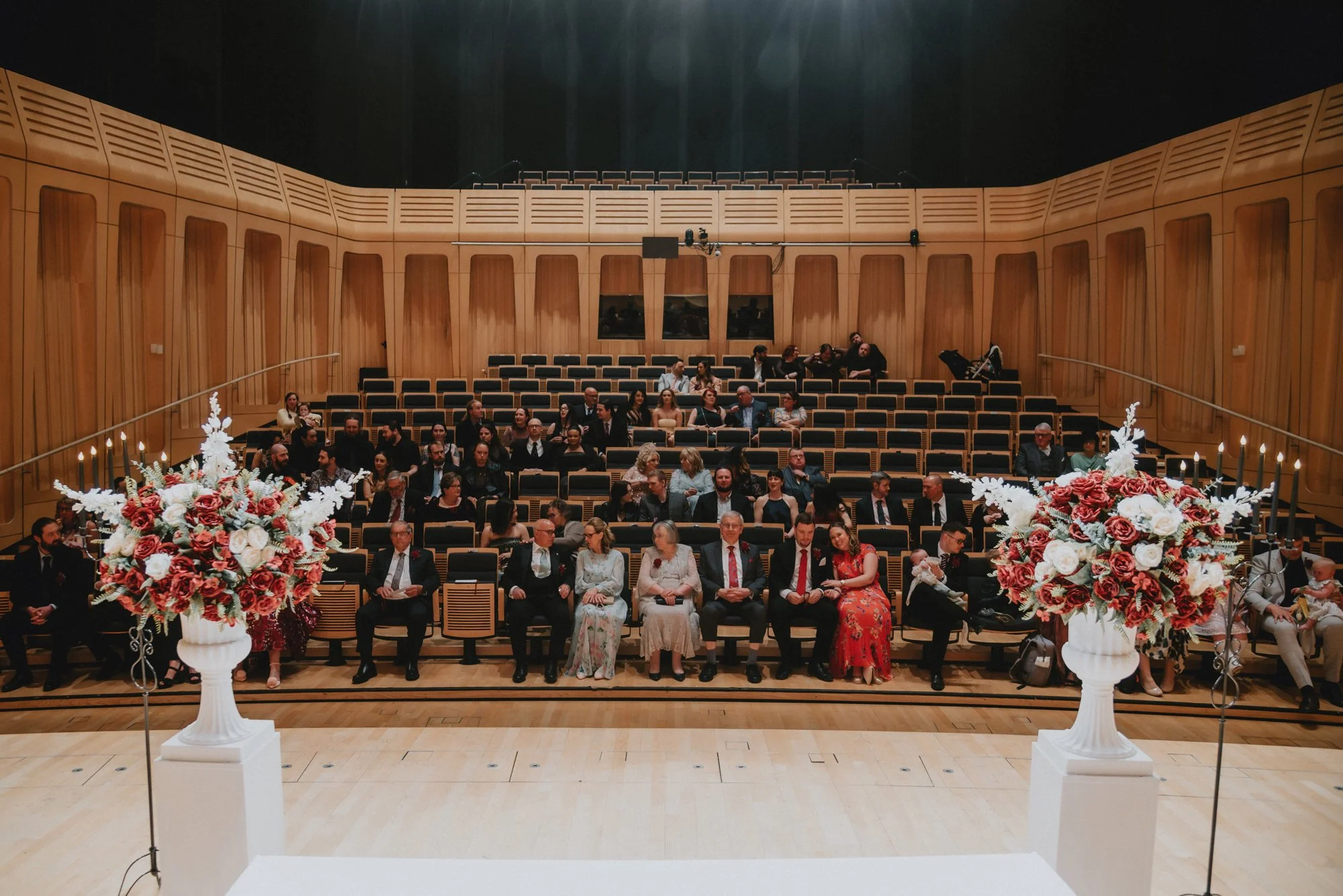 An audience seated in a concert hall with wooden walls, two large floral arrangements with pink and white flowers, and tall candle holders in the foreground.
