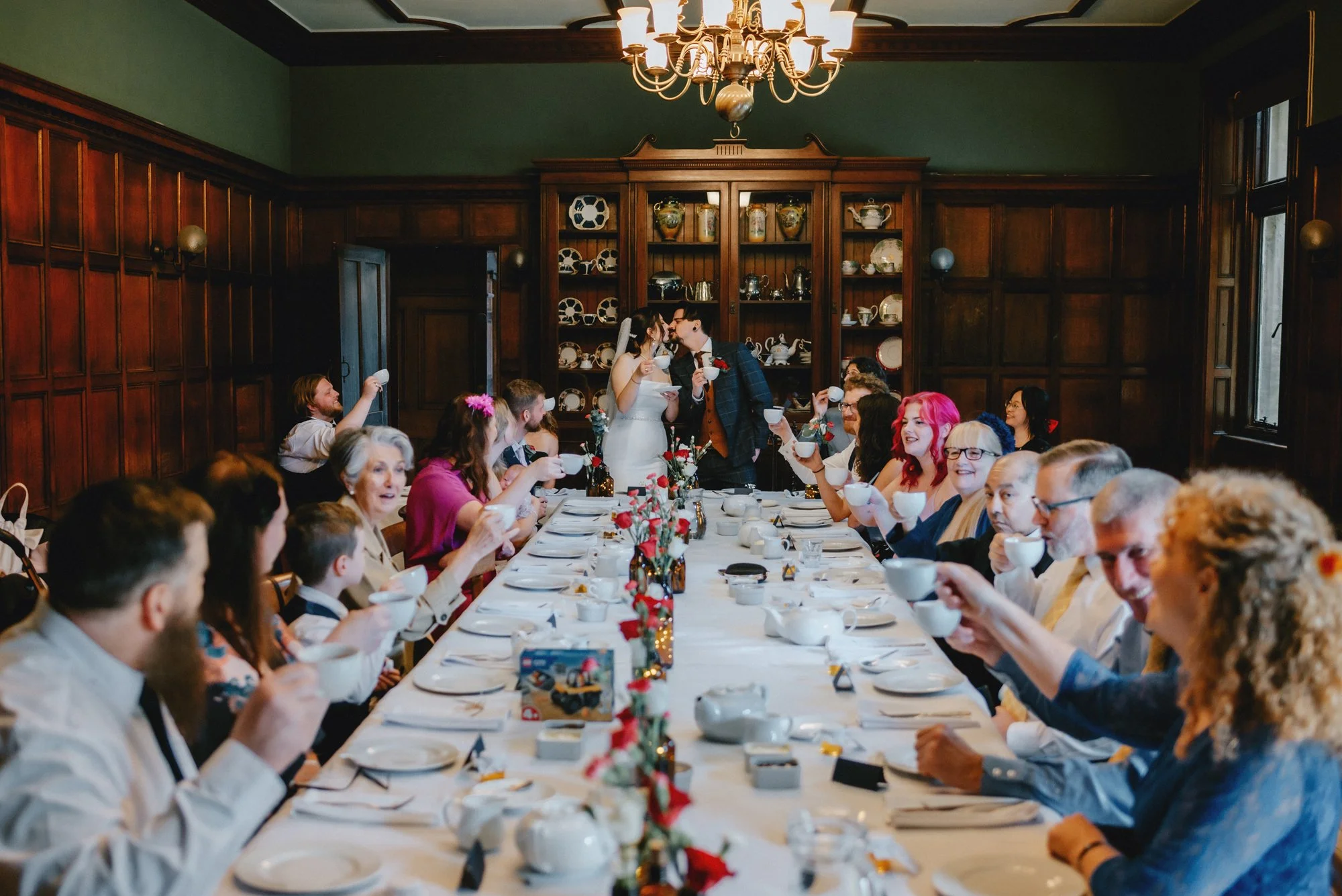 A wedding reception with a long dining table filled with guests raising teacups in a toast.