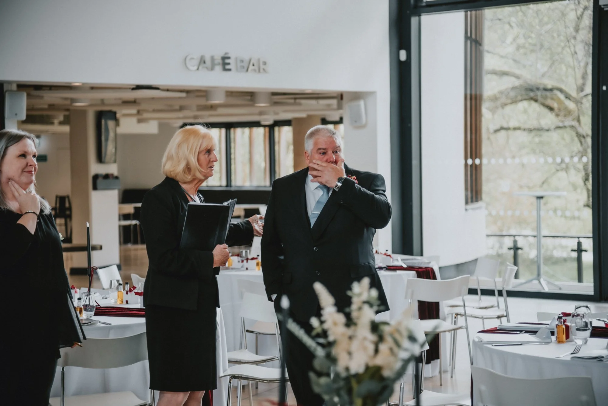 People in formal attire at a restaurant or event space, talking, with a sign that says 'Café Bar' in the background.
