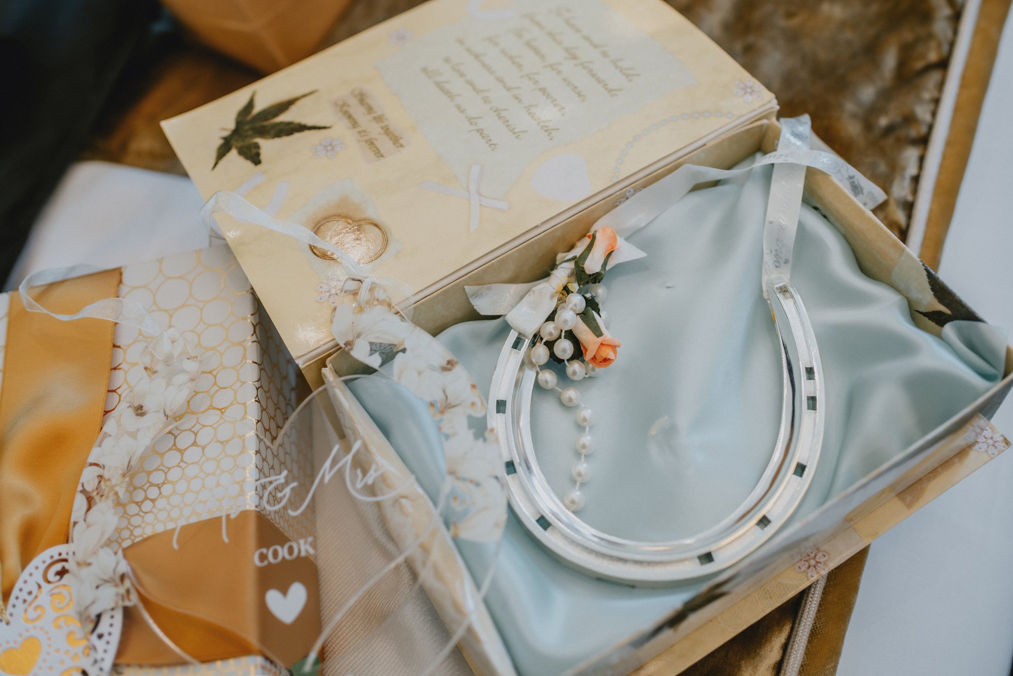 A gift or keepsake box containing a horseshoe, a pearl necklace, and a small bundle of flowers, including a peach-colored rose, placed on a satin fabric. There are additional items, including a decorative card or invitation, partially visible around 