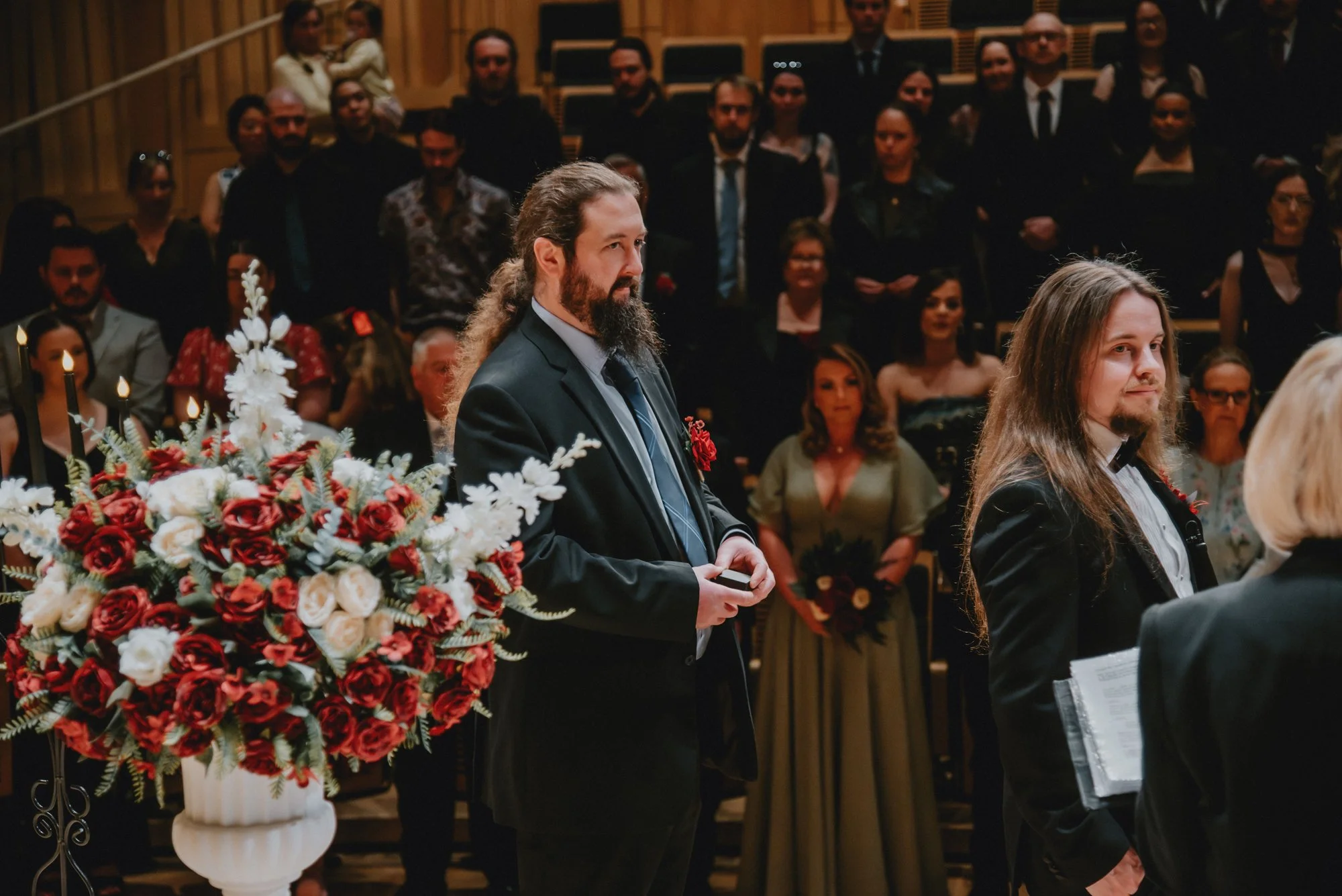 People participating in a formal ceremony or wedding, with two men in suits at the front, one holding a small object, and a large floral arrangement nearby, with an audience in the background.