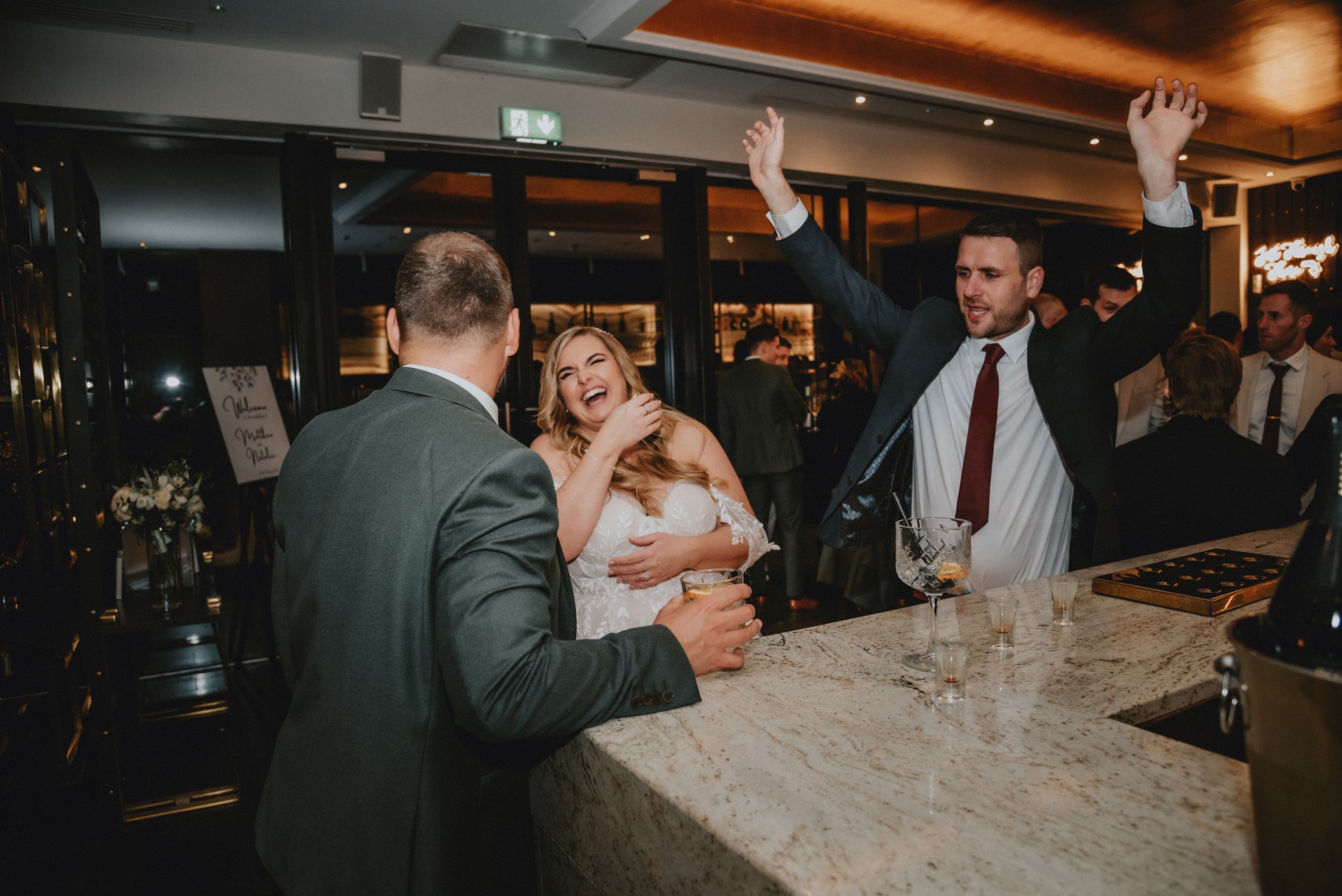 People laughing and talking at a bar during a celebration, with a woman in a white dress, men in suits, and drinks on the bar counter.
