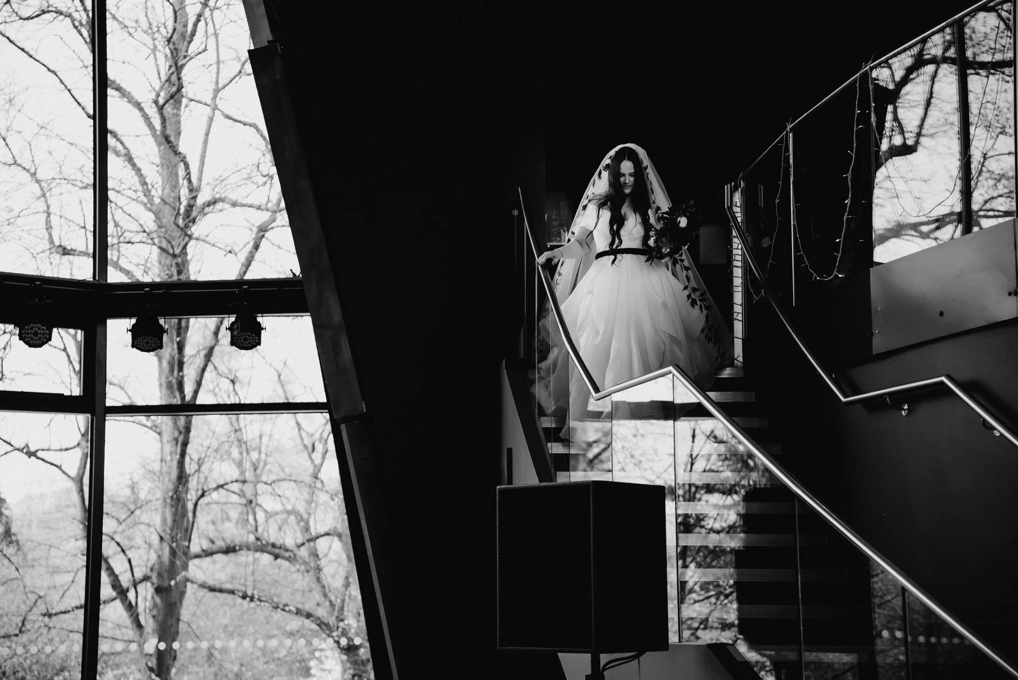 A woman in a wedding dress walking down a staircase inside a building with large windows showing trees outside.