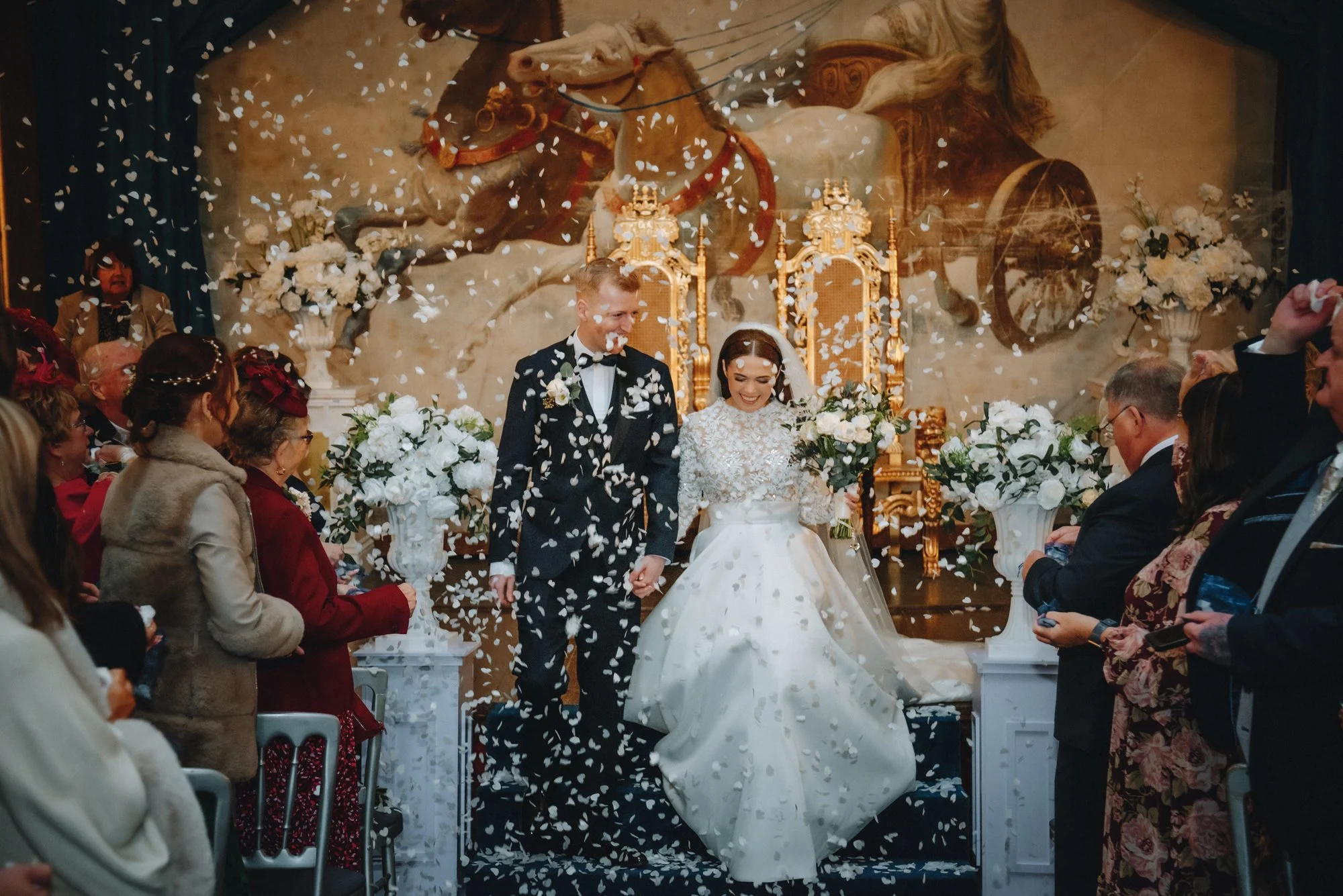 A wedding ceremony with a bride and groom walking down the aisle, surrounded by guests. The bride is wearing a white wedding dress and holding a bouquet. The groom is in a dark suit with a bow tie. White petals are falling, and the background feature