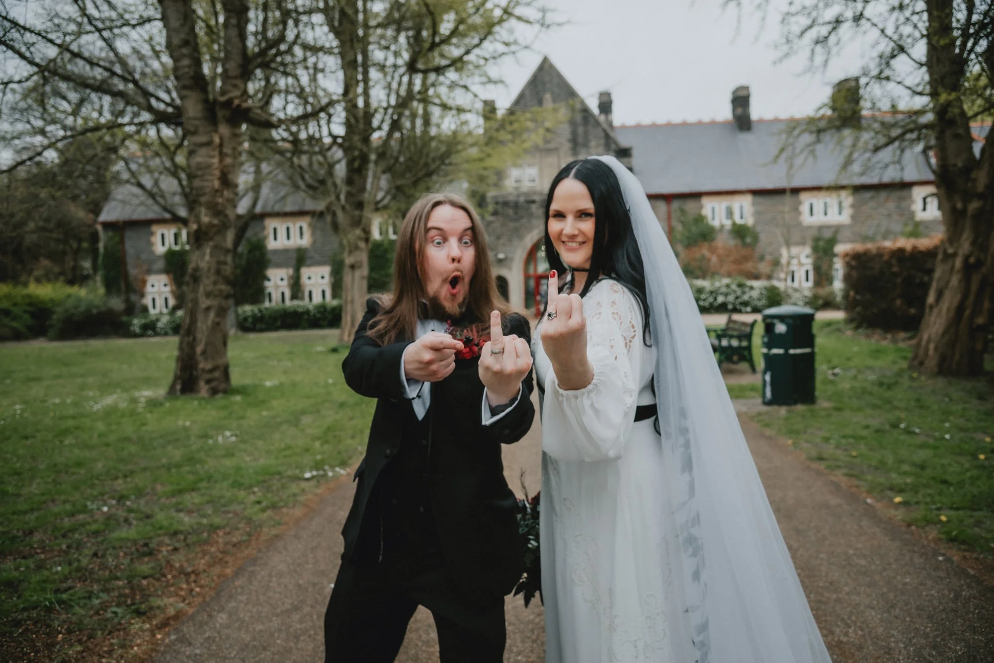 A bride with long black hair in a white wedding dress and veil, standing next to a groom with long brown hair and a beard, in a black tuxedo, both showing their middle fingers in a playful, rebellious pose on a path in a park with trees and a large s