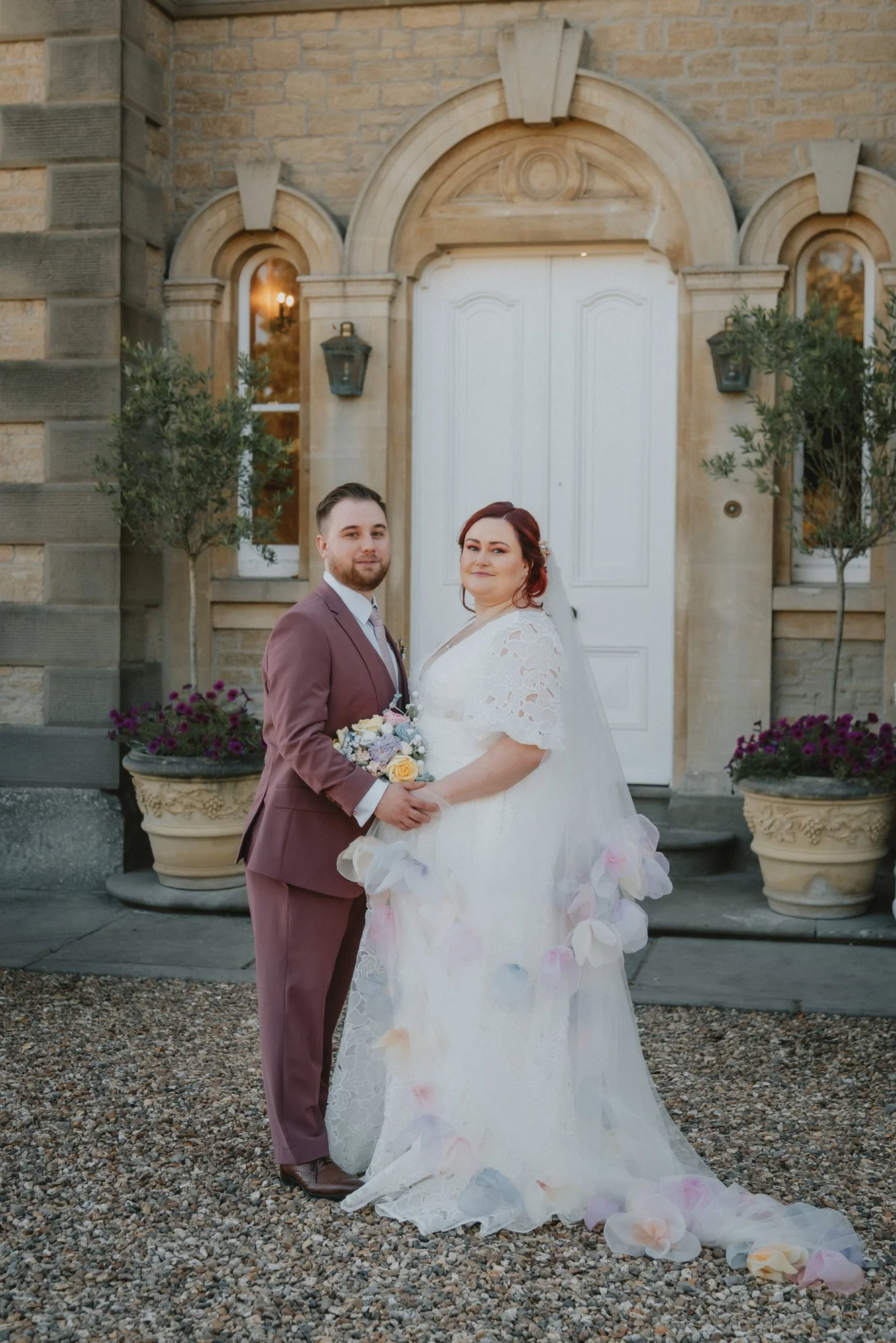 A newlywed couple standing outside a brick building, holding hands and smiling. The bride wears a white lace wedding dress with petal details, and the groom is in a mauve suit with a white shirt and light tie. They are in front of a white door with a