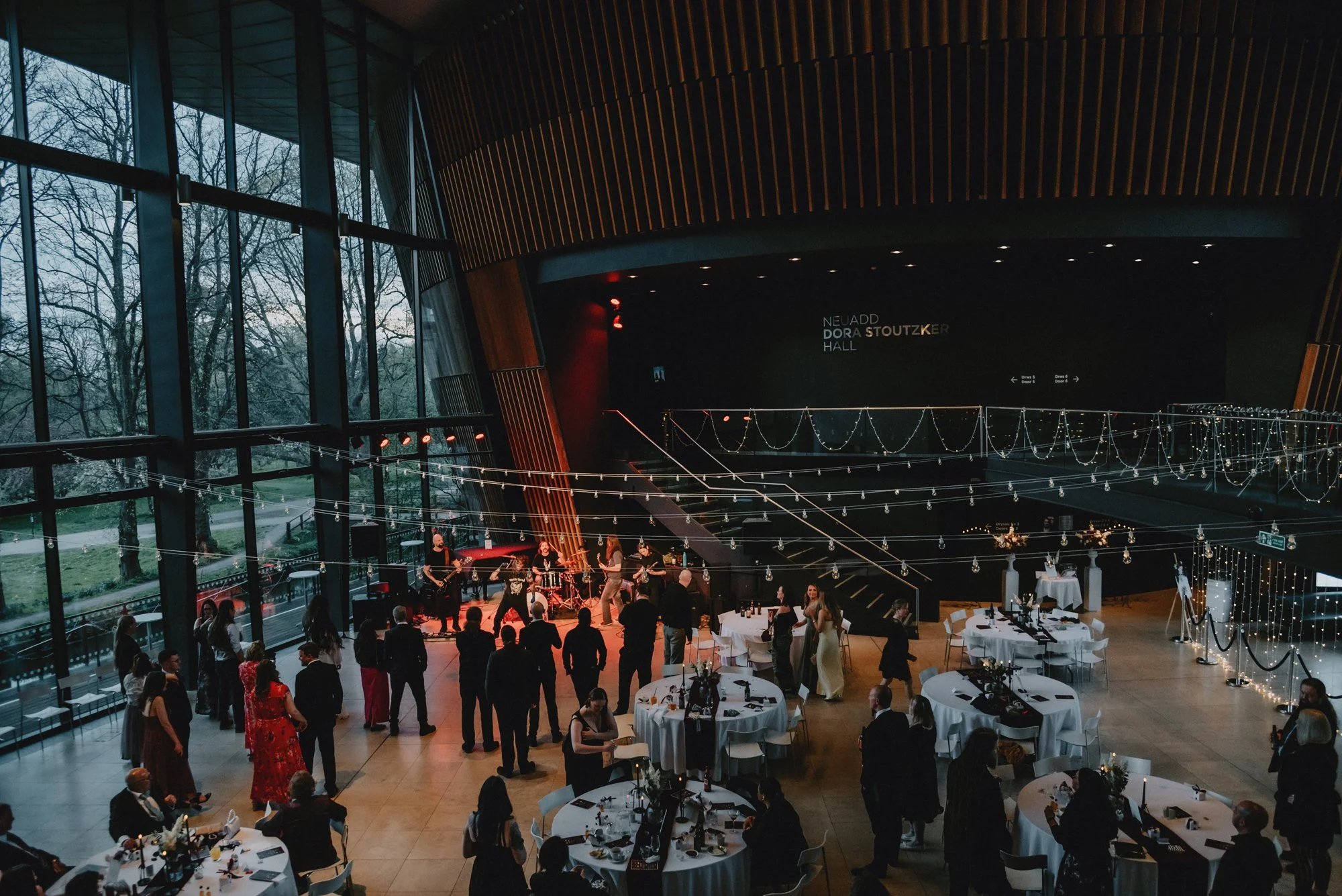 People attending a concert or event in a modern, large hall with string lights, tables, and a band performing on stage.